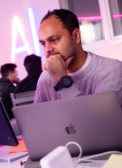 A man sits at a desk looking at a computer. A neon AI sign is visible in the background.