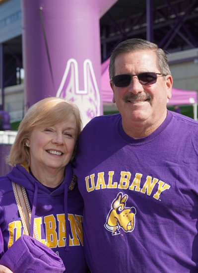Two people wearing UAlbany branded purple t-shirts stand at an outdoor event.