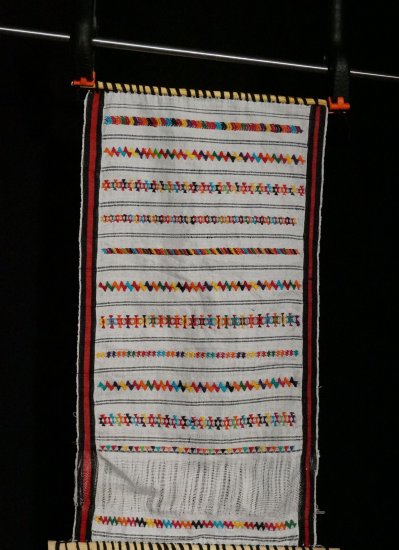 A red and white weaving with multicolored embellishments on a small loom.