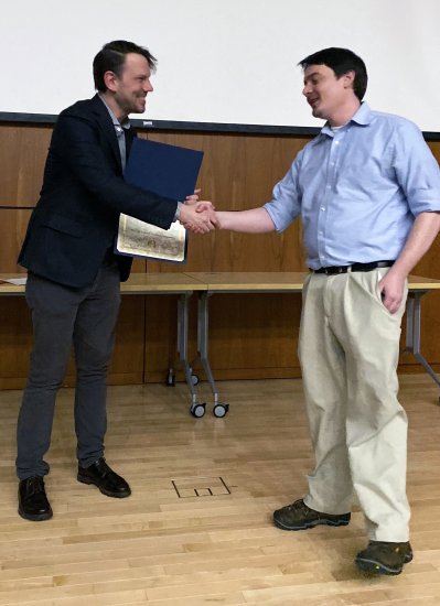 A man in a blue shirt and khakis shakes hands and accepts an award certificate from a man in a suit jacket.