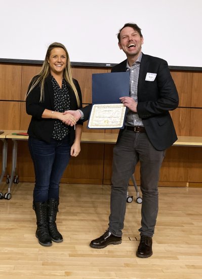 A woman in a patterned top, black cardigan and jeans shakes hands with accepts an award certificate from a man in a suit jacket.