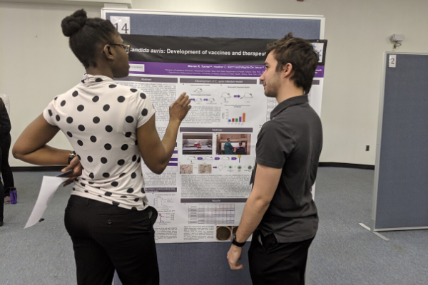 Two students talk in front of a research poster. One student wears a white shirt with black polka dots, and has her hand up as she explains the poster. The other student, wearing a grey shirt and black pants, stands and listens to the explanation of the poster.