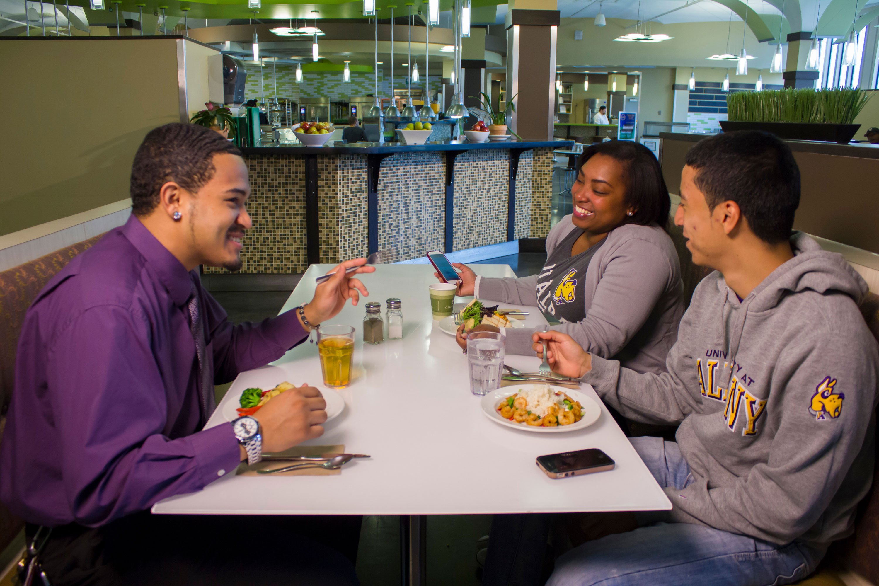 Three students sit together inside State Quad dining room, eating shrimp stir fry and Greek salad