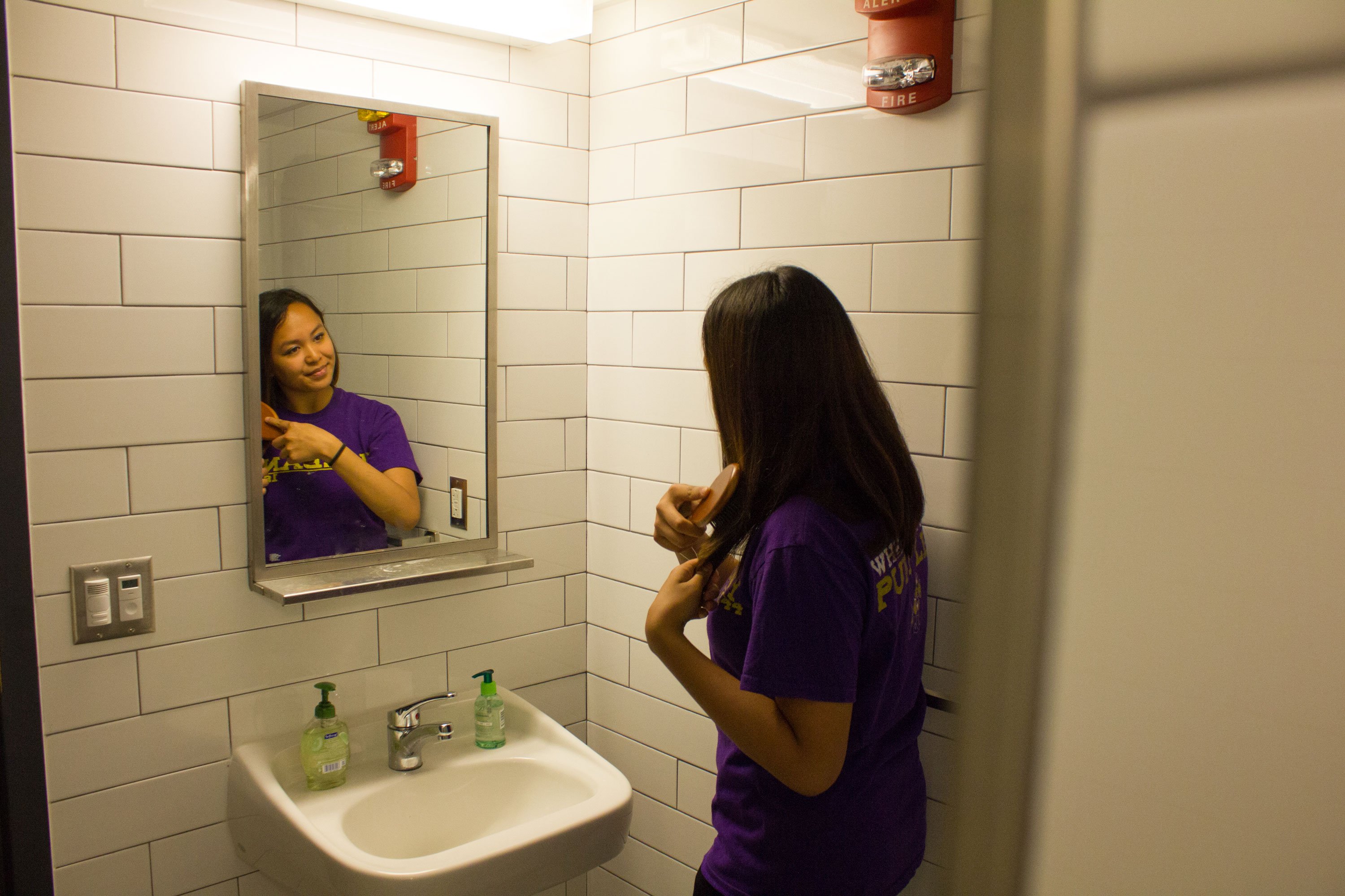 A student in a purple shirt brushes her hair in front of a bathroom sink and mirror