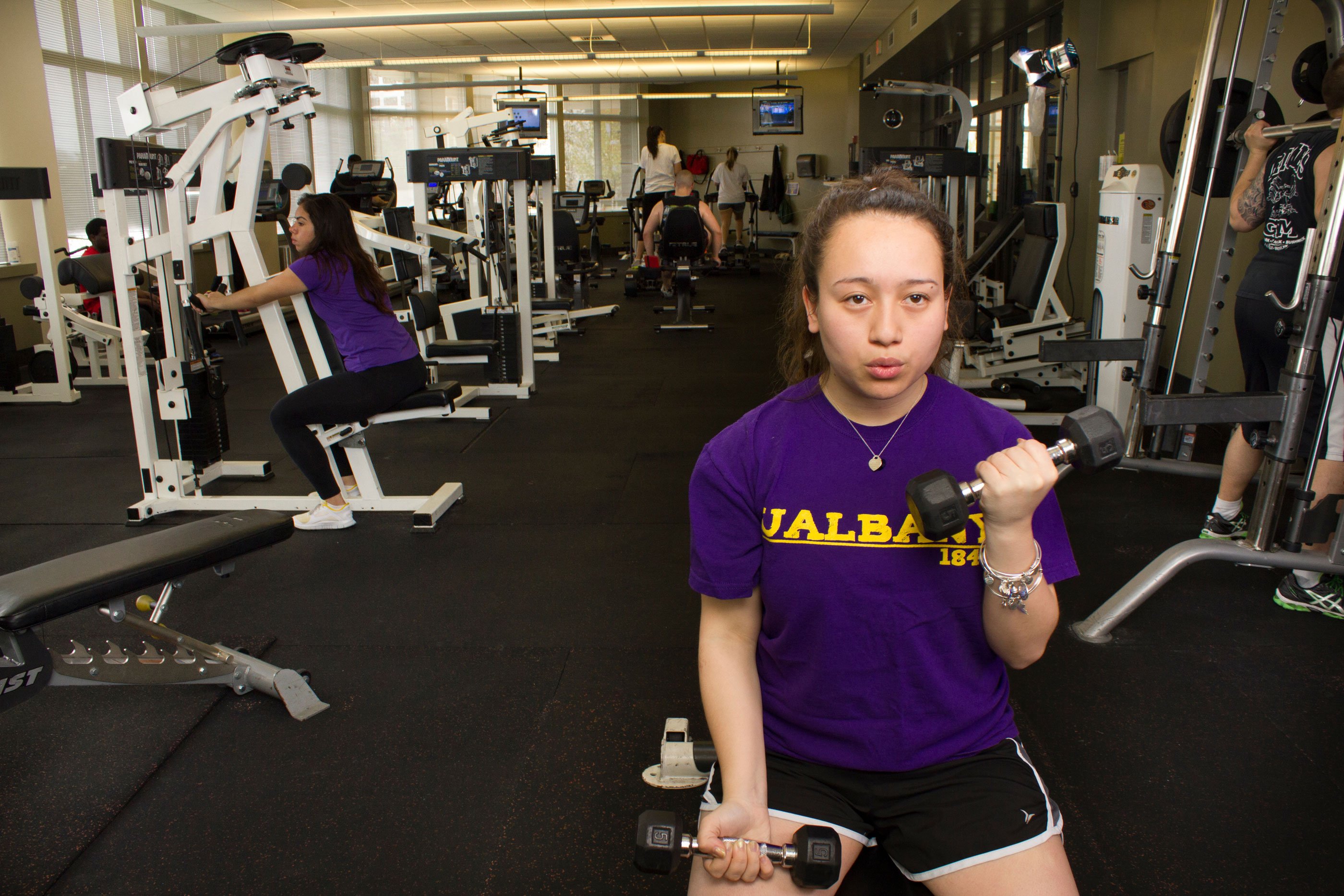 A student wearing a UAlbany shirt does bicep curls inside the Empire Commons fitness center