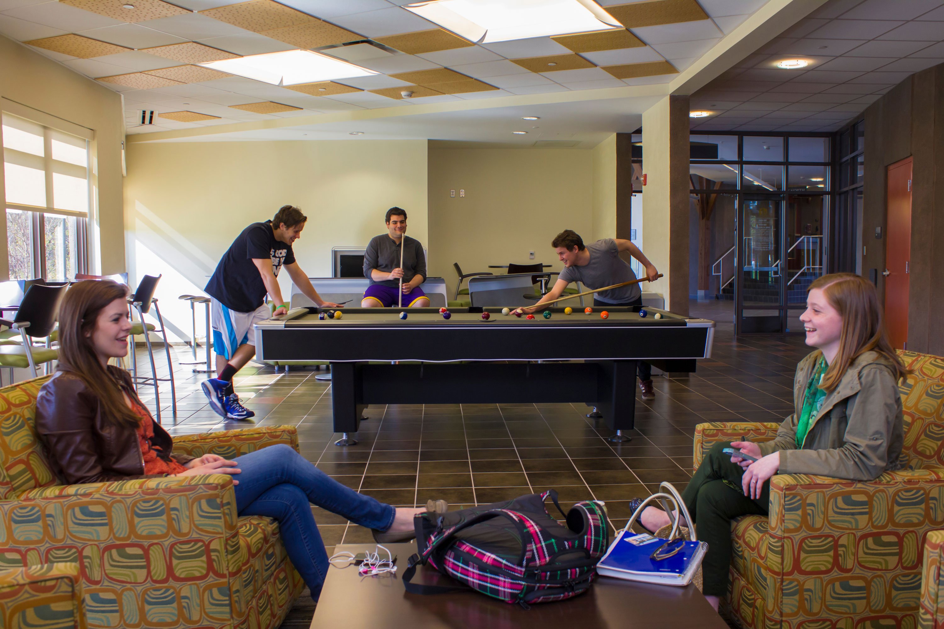 Two students sit and talk, while three others place billiards, inside a Liberty Terrace lounge