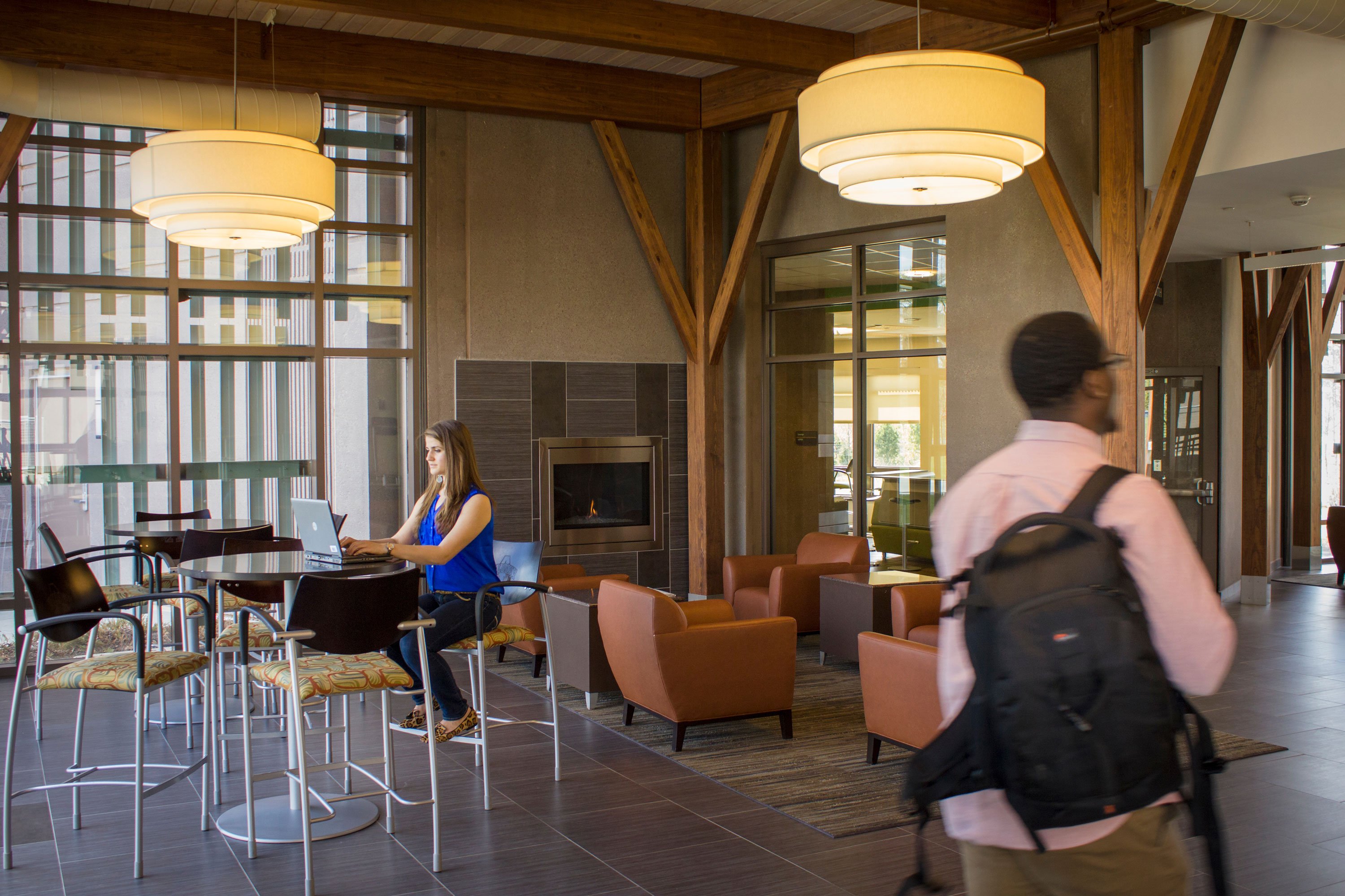 A student works on her laptop, while another walks by, in the Liberty Terrace lobby, which has a fireplace, high top tables and arm chairs