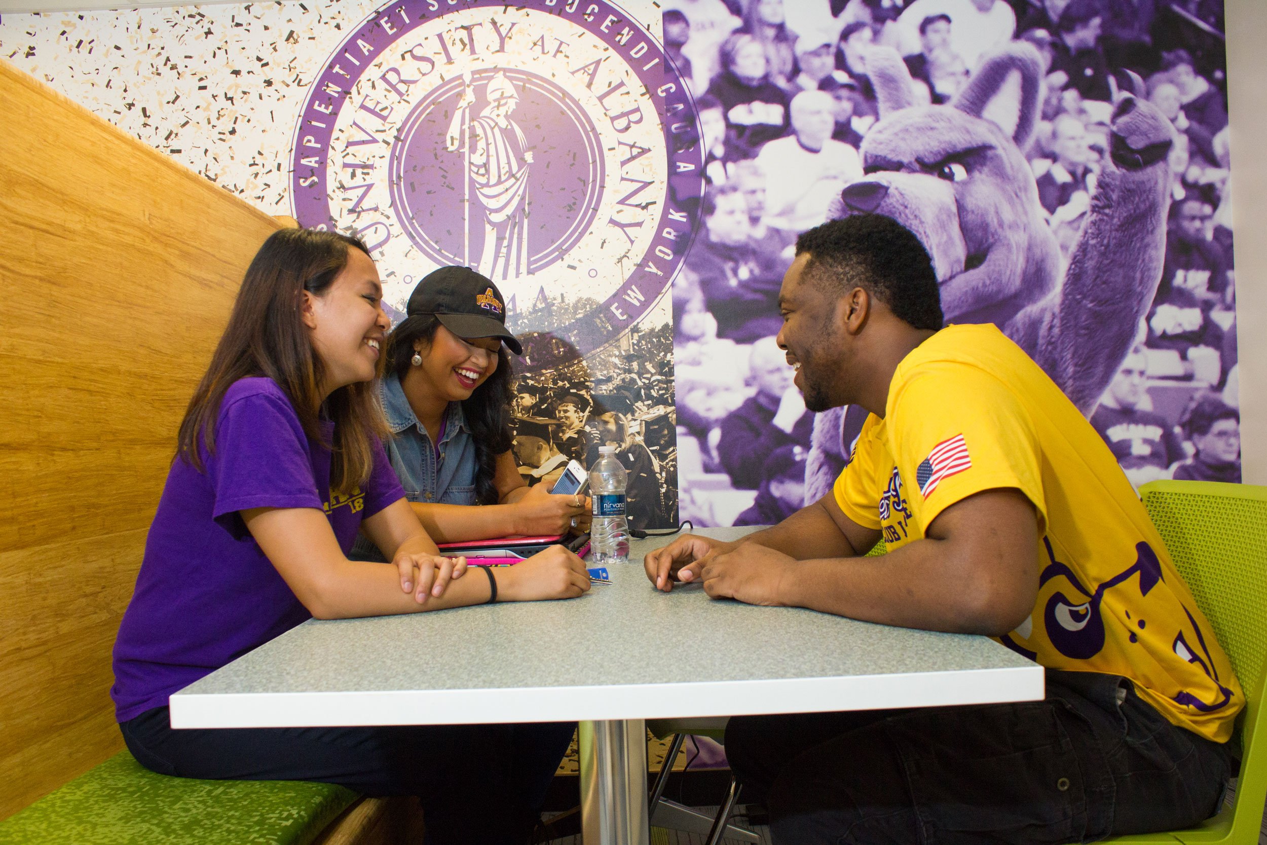 Three laughing students sit at a table with a green chair and bench, with the UAlbany seal on the wall