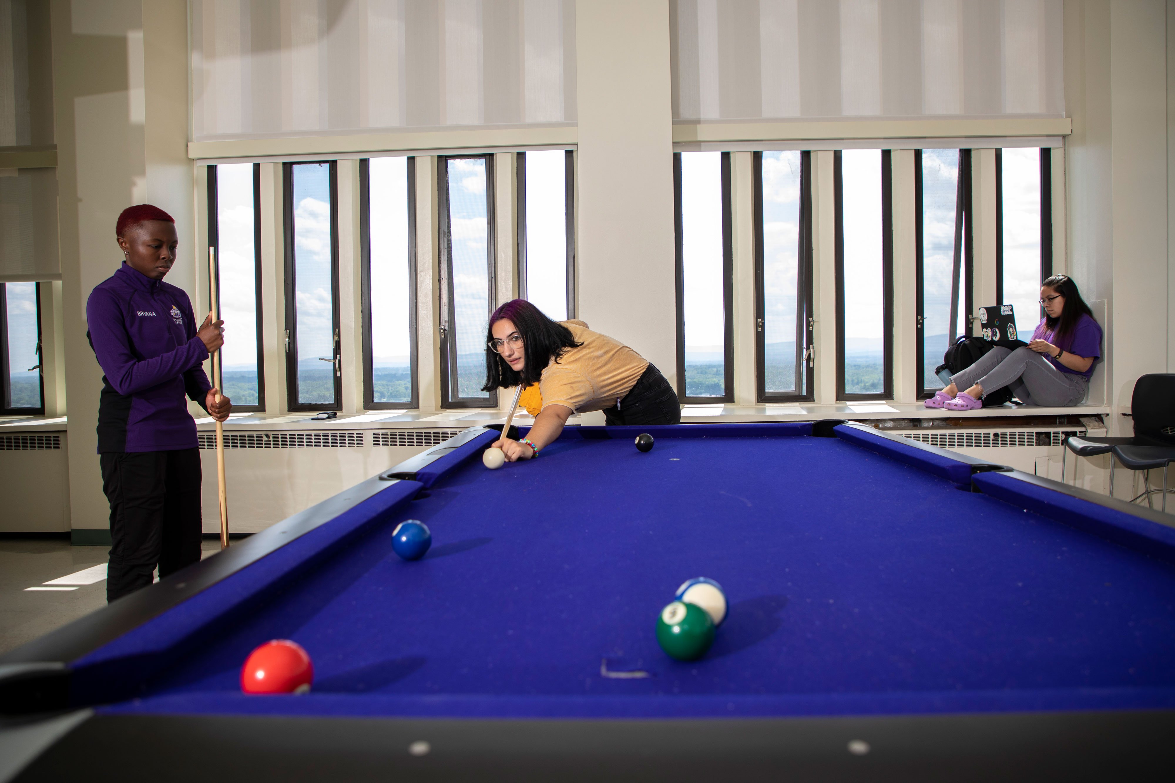Two students play billiards on a blue pool table, while another student works on her laptop while perched on a window sill overlooking a view of Albany