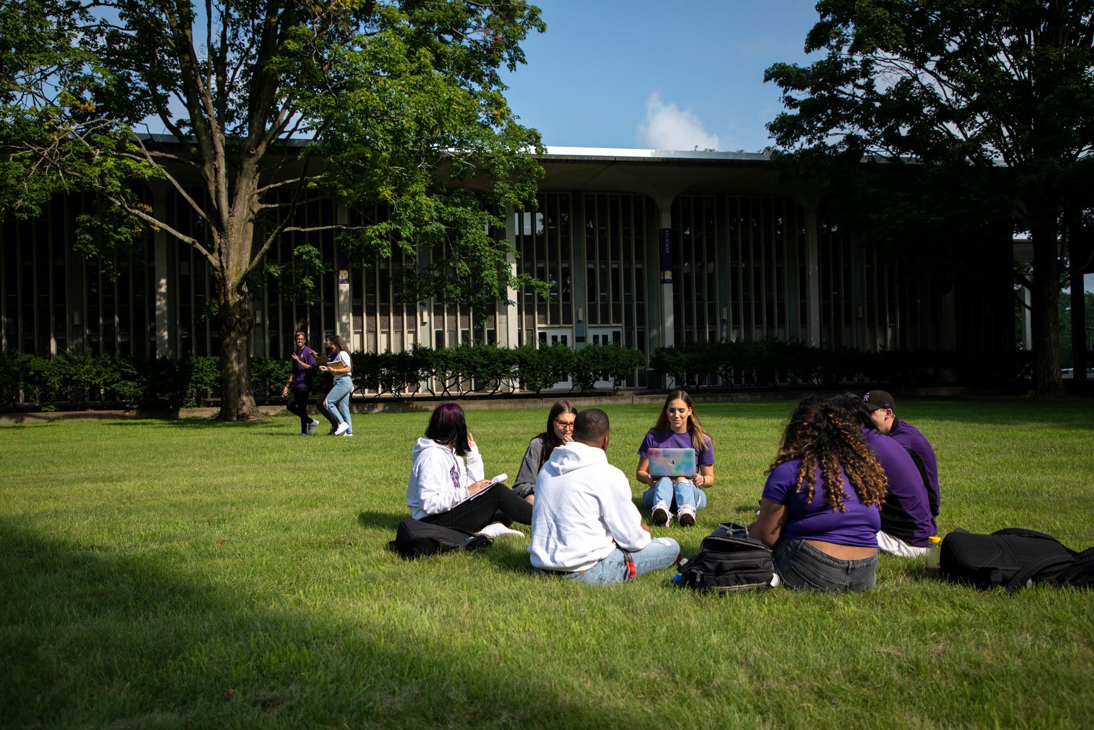 Seven students sit in a circle on the grass outside Dutch Quad, while two other students walk by on a sunny day
