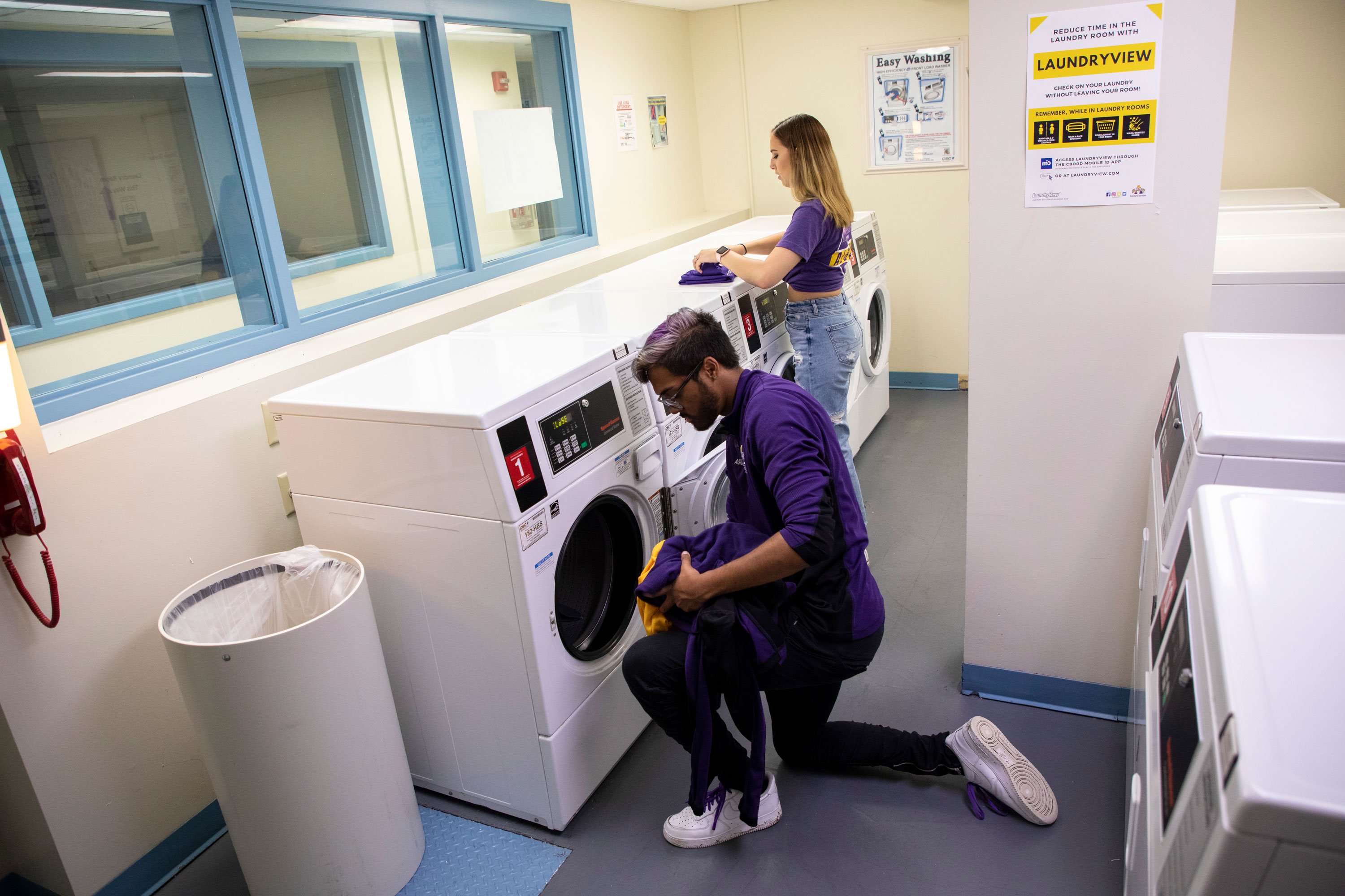 One student unloads a laundry machine, while another folds clothing, inside a laundry room