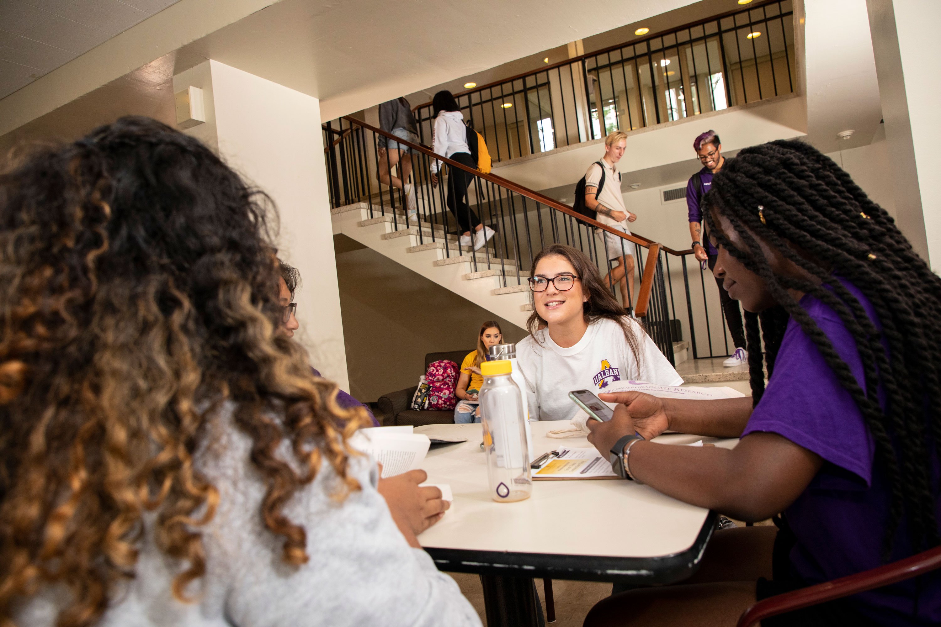 A student smiles as she talks to three other friends seated at a table with her. Other students walk through the residence hall and up its stairs.