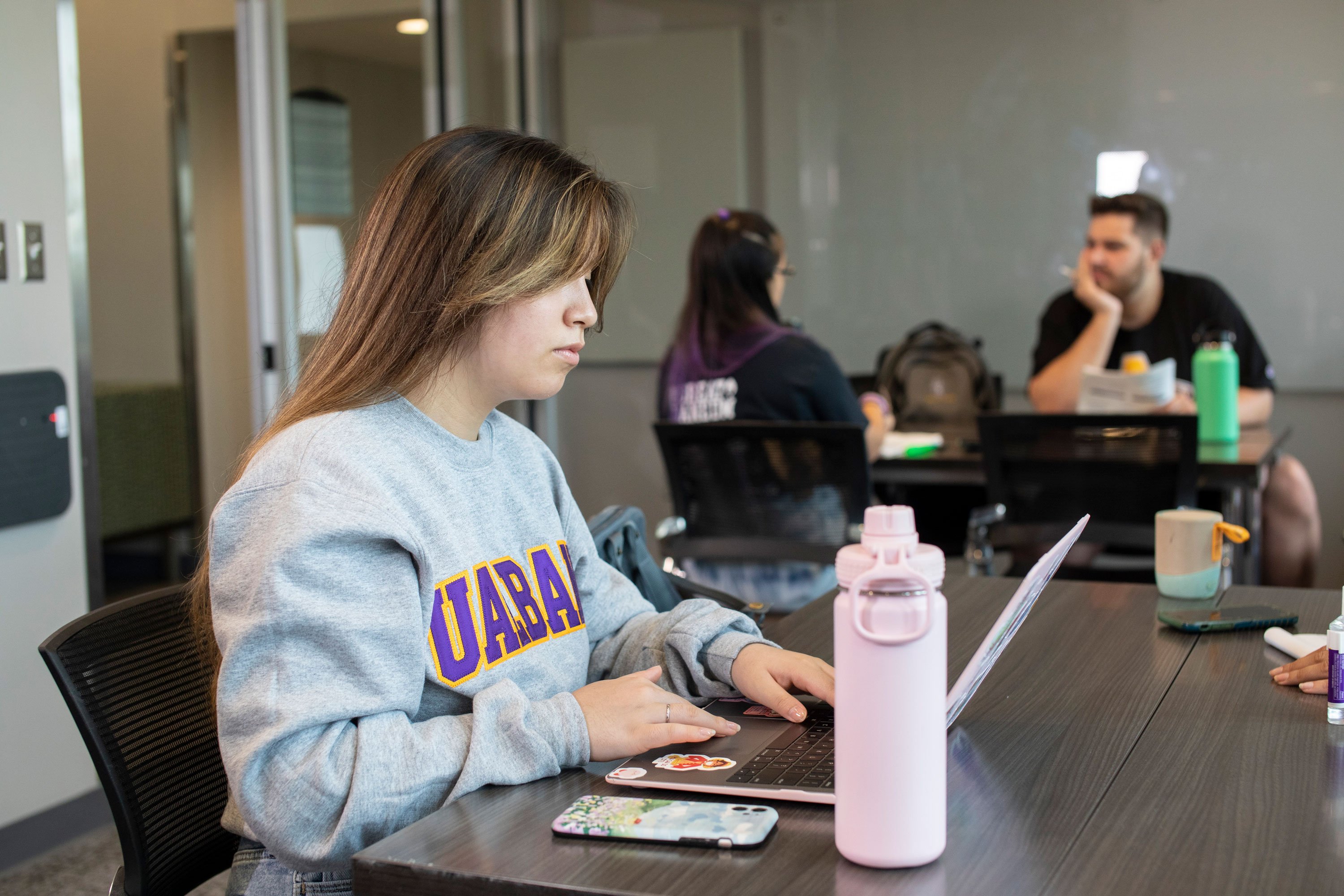 A student works on her laptop, with a phone and water bottle close by, while two other students talk in the background, inside a study room