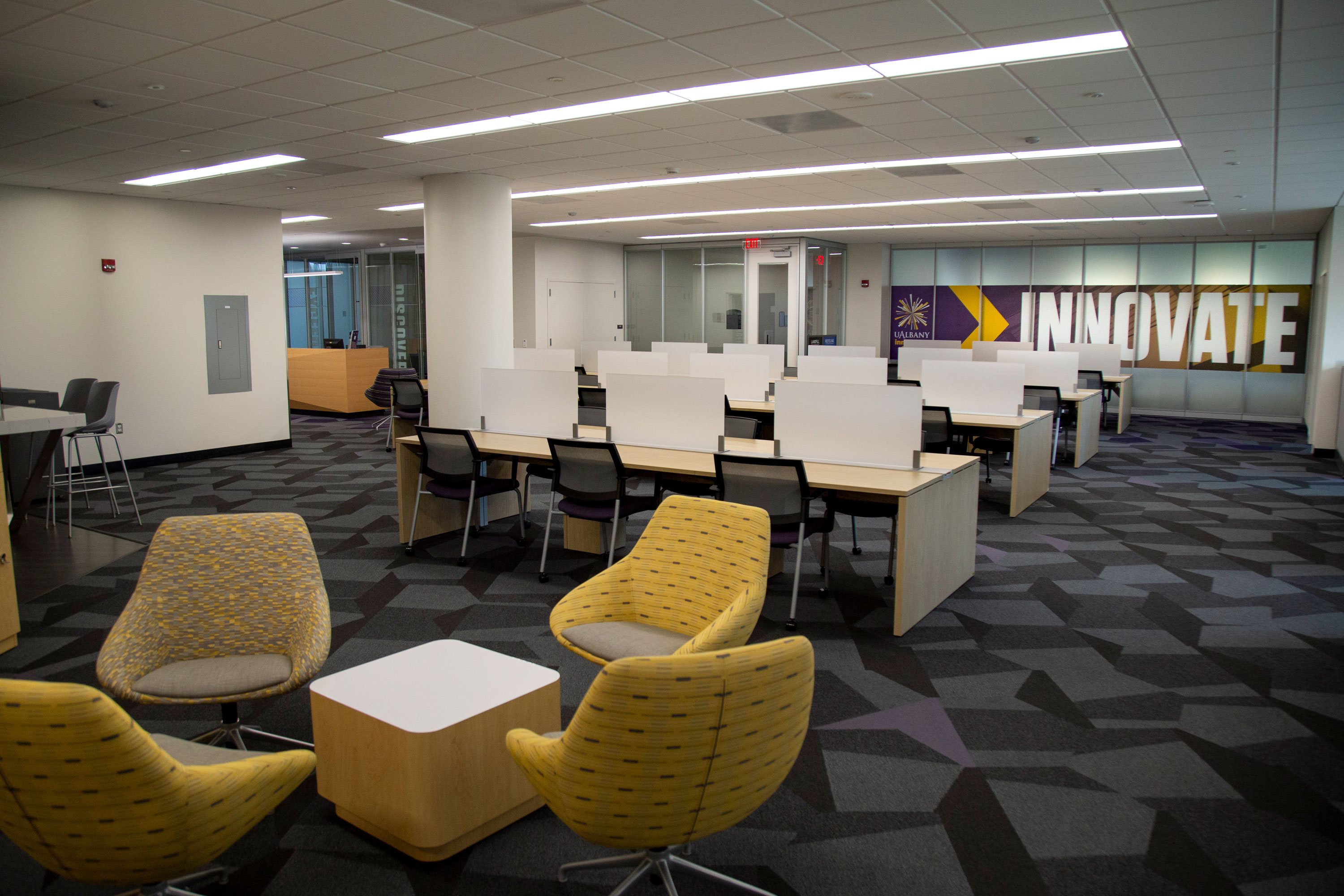The inside of the UAlbany Innovation Center, with four yellow chairs around a coffee table and about 40 shared workstations situated in front of the Innovate sign.