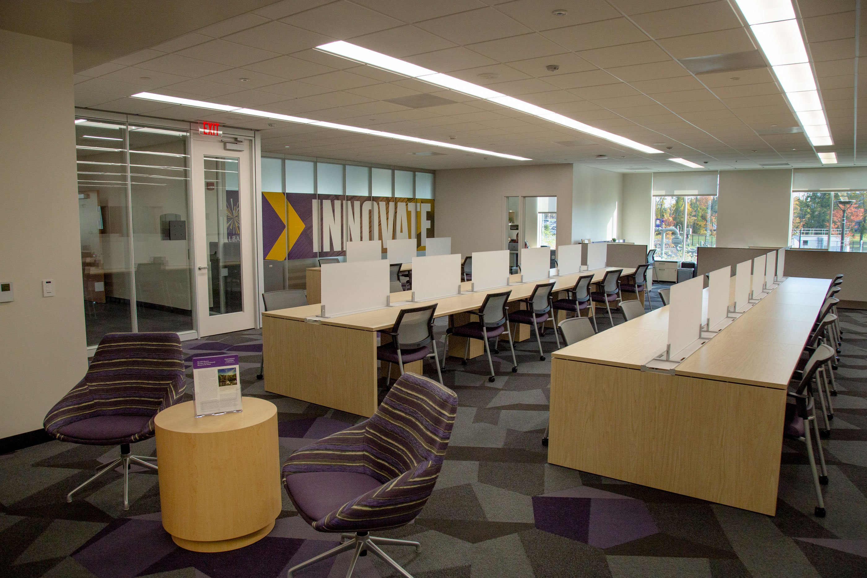 The inside of the UAlbany Innovation Center, with two purple chairs around a coffee table and about 40 shared workstations situated in front of the Innovate sign.