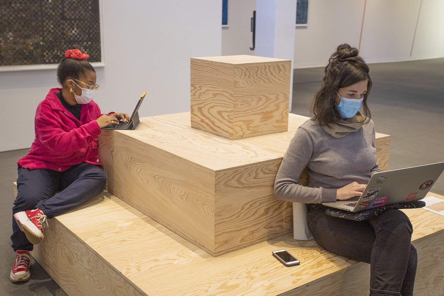 Two visitors working on their laptops sitting on a plywood pyramid sculpture