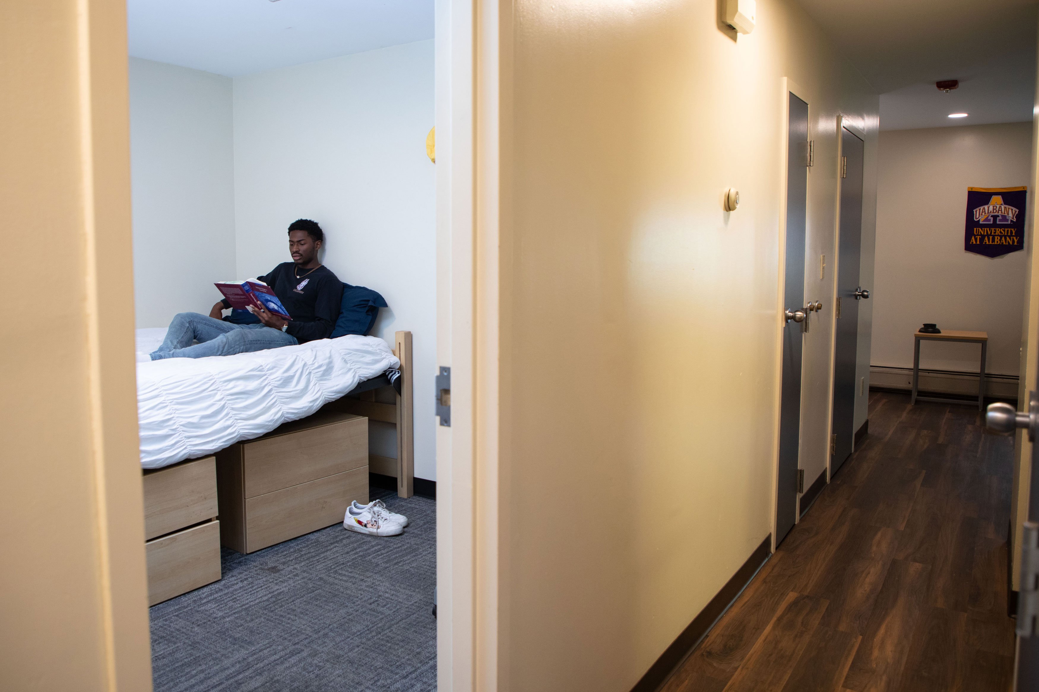 A view down an apartment hallway and into a single room. A student sits on his bed reading.