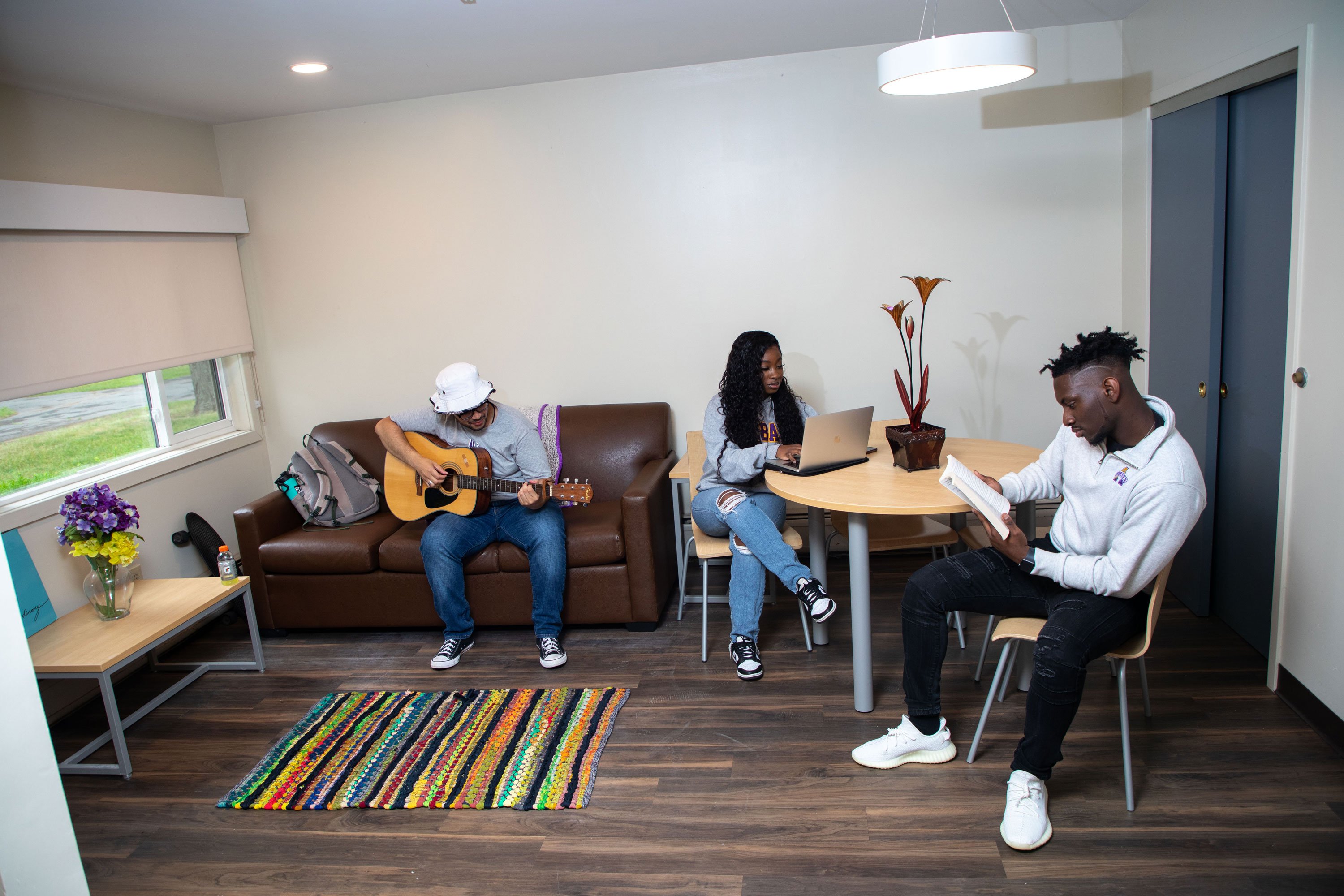 Three students sit inside a living room with a four-seater table and a brown couch. One student plays guitar, another works on a computer and a third reads a book.