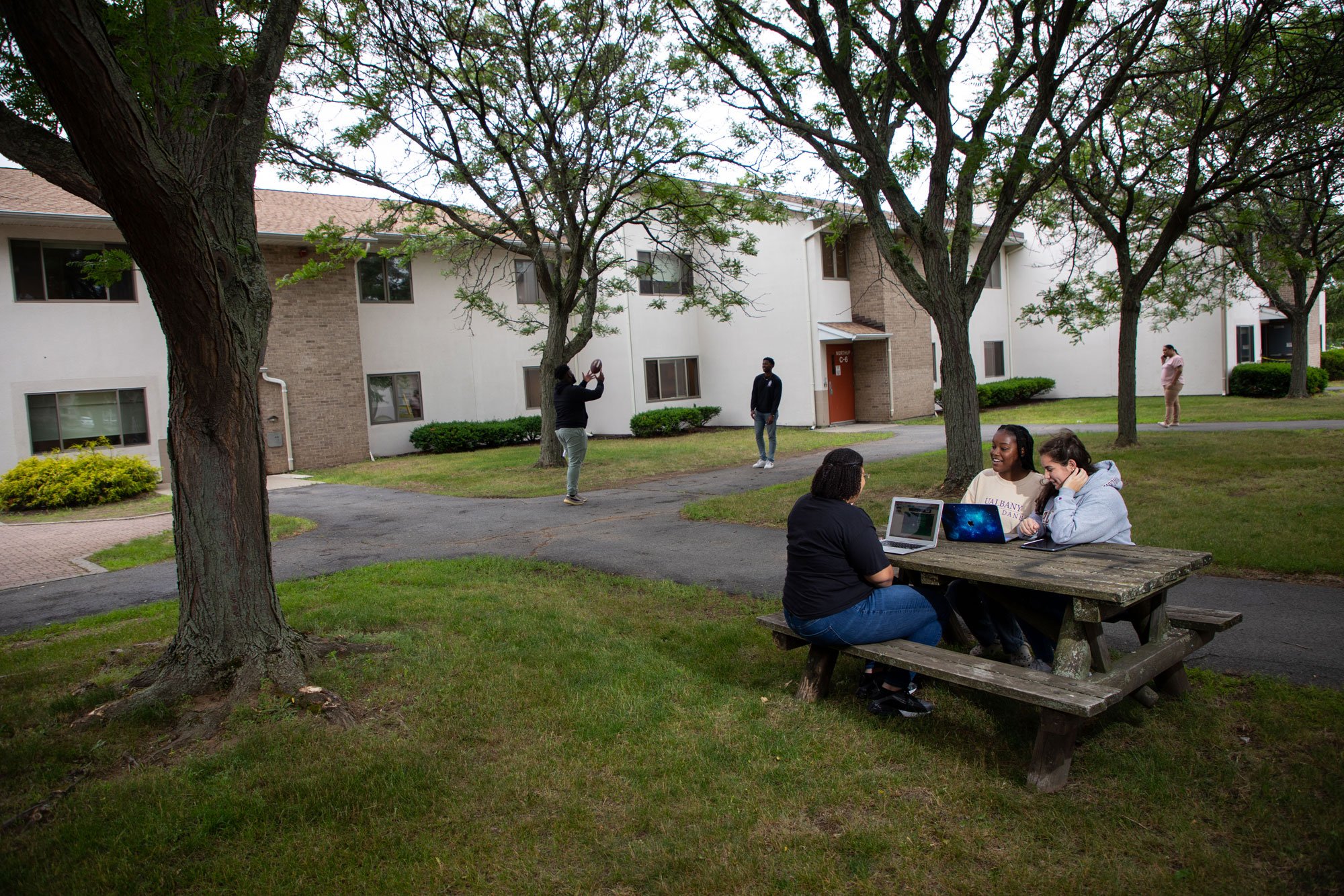Three students sit with laptops at a picnic table outside Freedom Apartments, while two other students play catch with a football behind them.