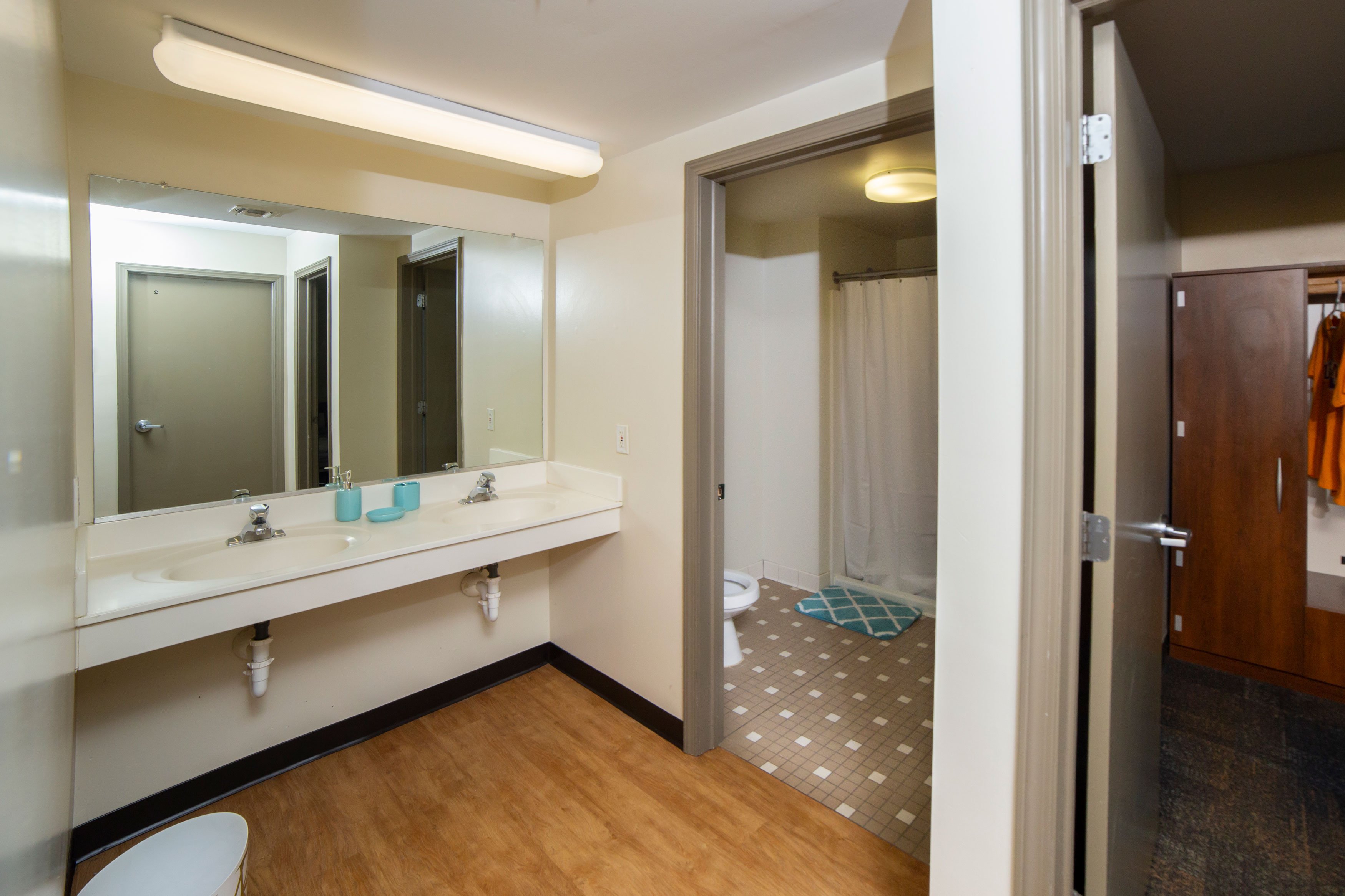 A two room bathroom, with a double sink in one room and a toilet and shower in the other adjoining room.