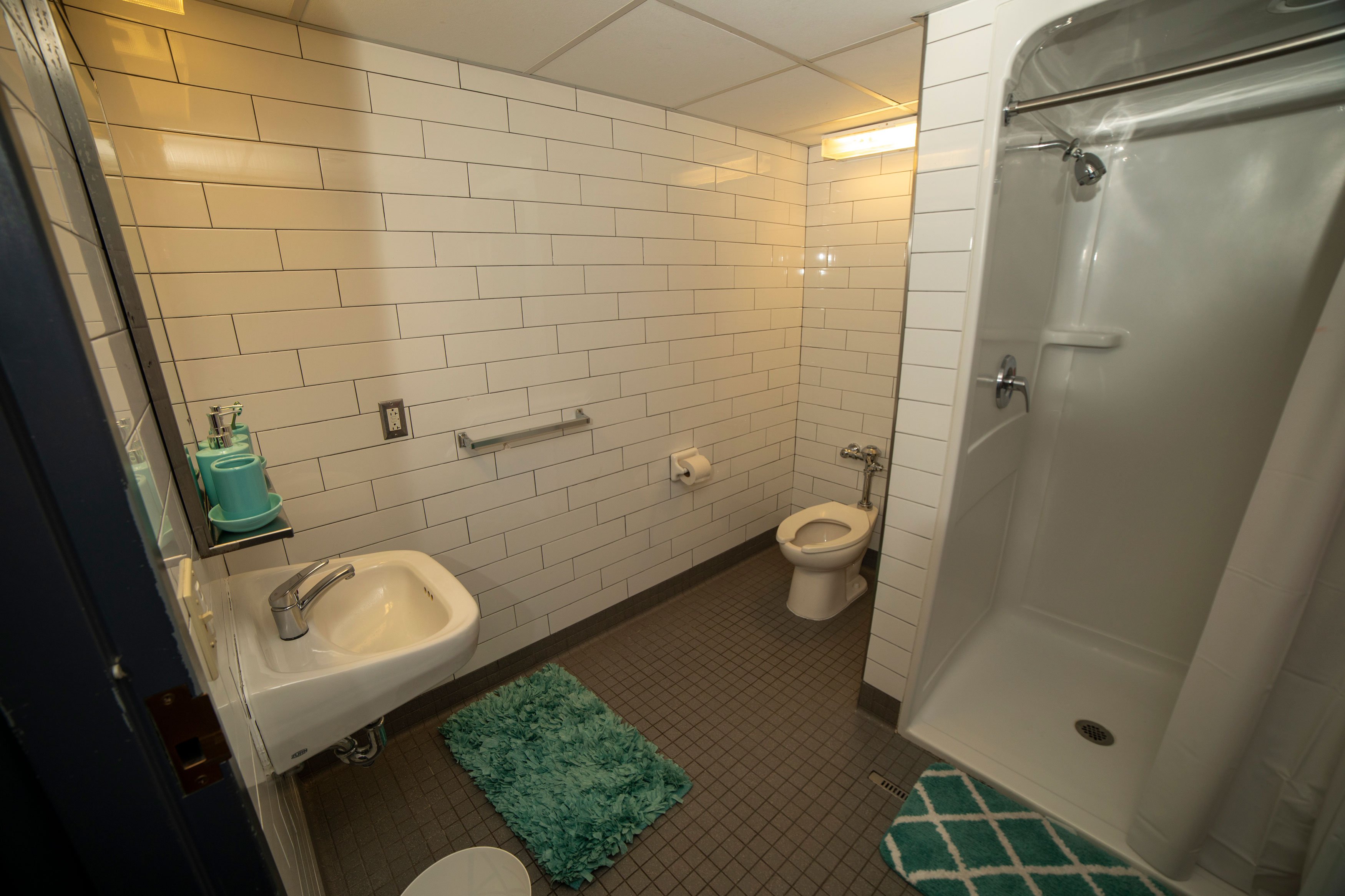 A bathroom with a walk-in shower, toilet and sink. The wall tiles and fixtures are white, the floor tiles are brown and there are turquoise bath maps and soap dispensers.