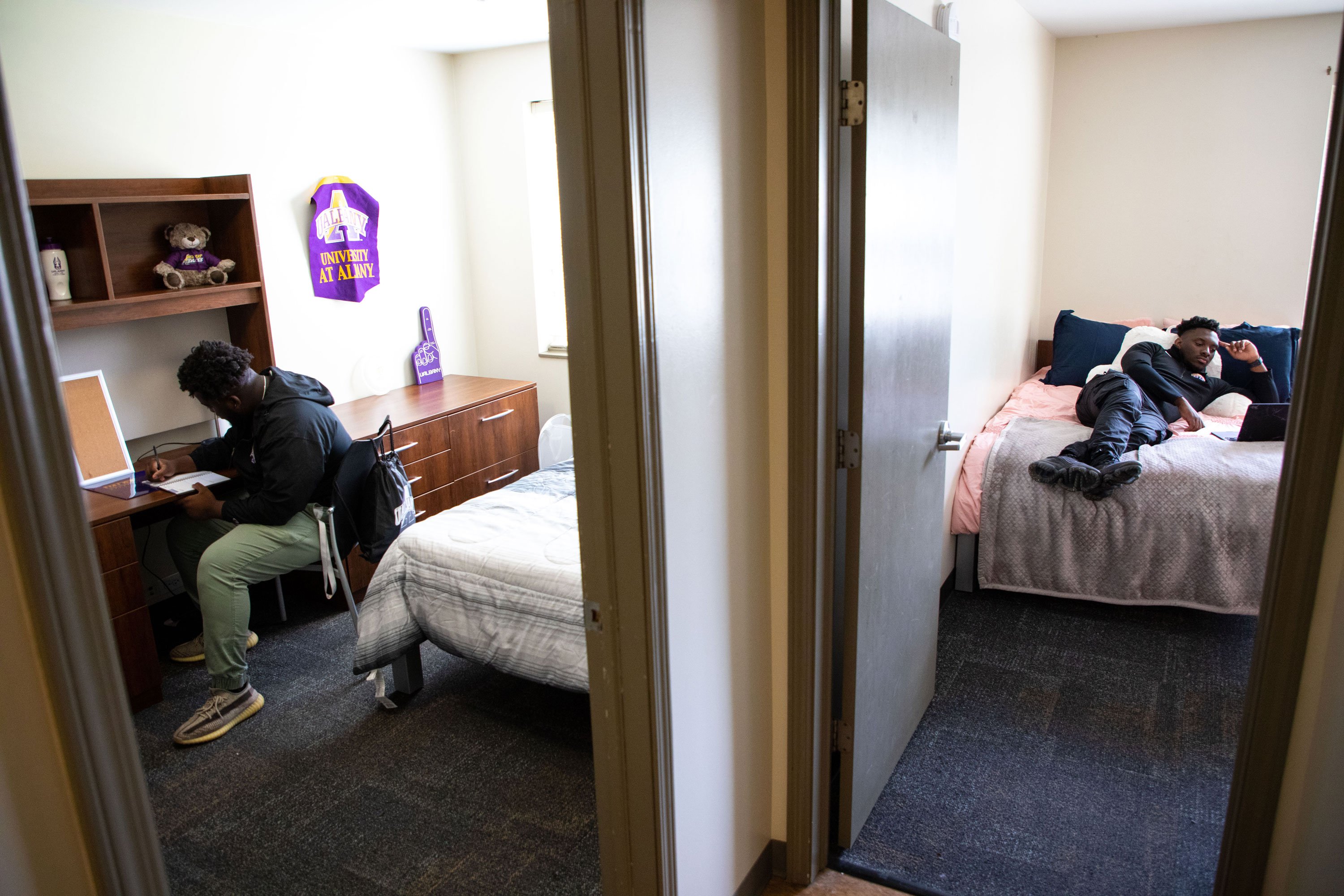 Two doors open into separate bedrooms. In the bedroom to the left, a student sits at a desk and writes in a notebook. In the bedroom to the right, a student lays on the bed and watches something on his laptop.