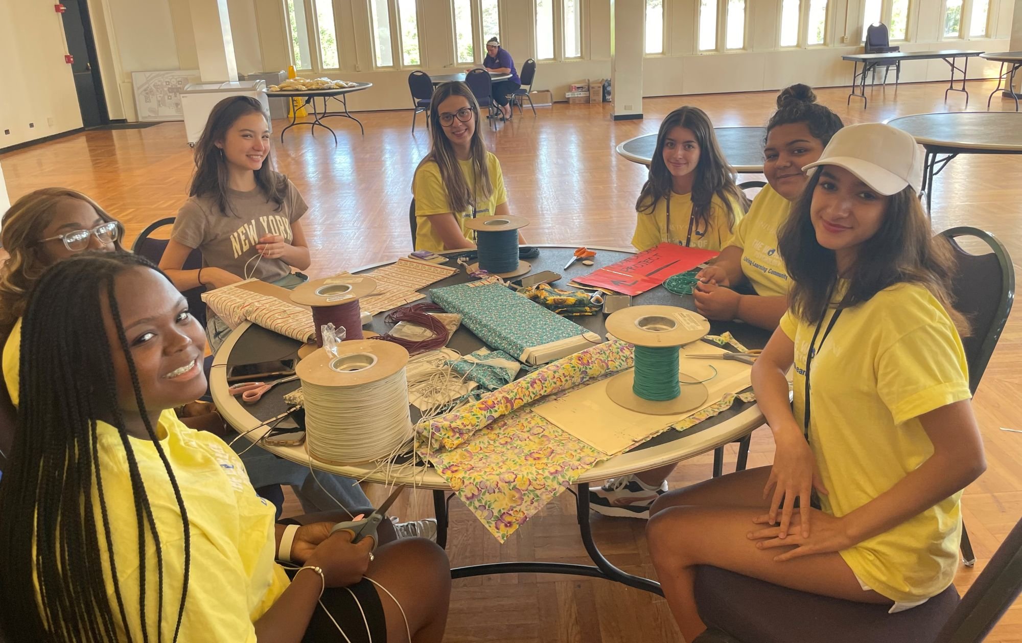 Seven UAlbany students sit around a craft table topped with patterned fabric and large spools of thread. Six are wearing matching yellow shirts.