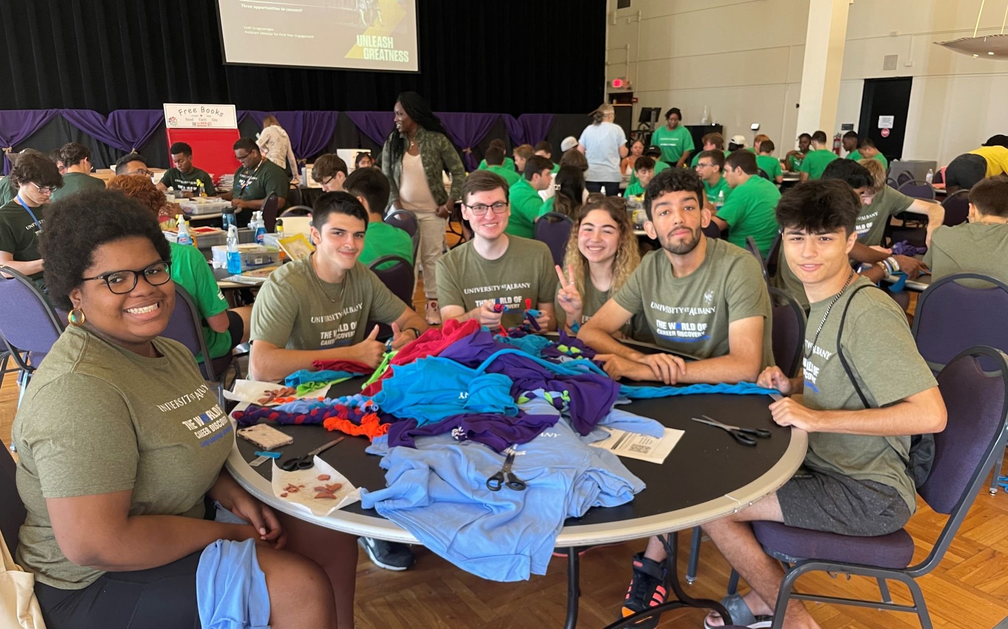 Six students in matching green T-shirts sit around a craft table and smile as they  pose for a photograph.