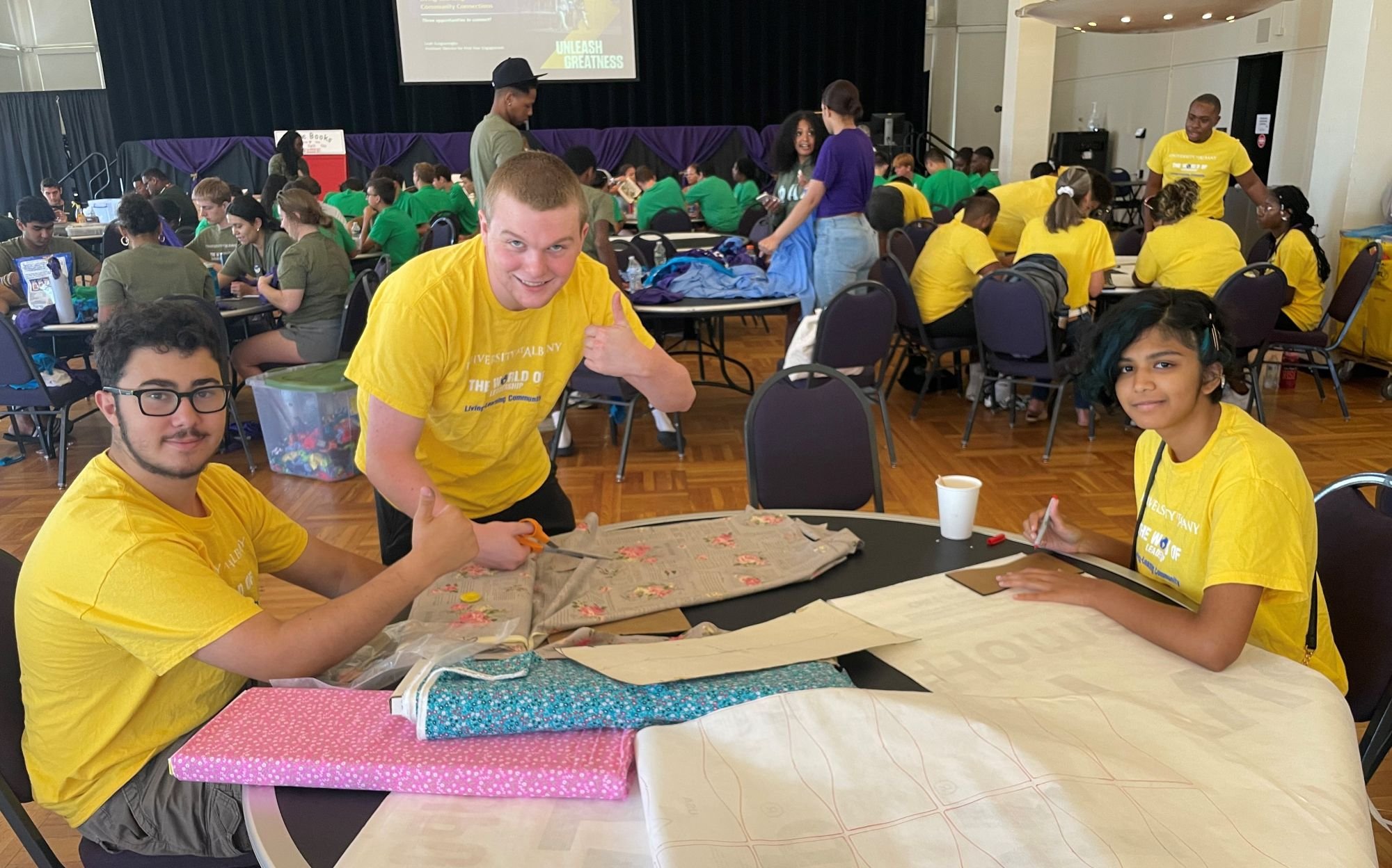Students in matching yellow T-shirts smile and pose for a photograph. Two of them are seated at a craft table topped with large pieces of fabric.