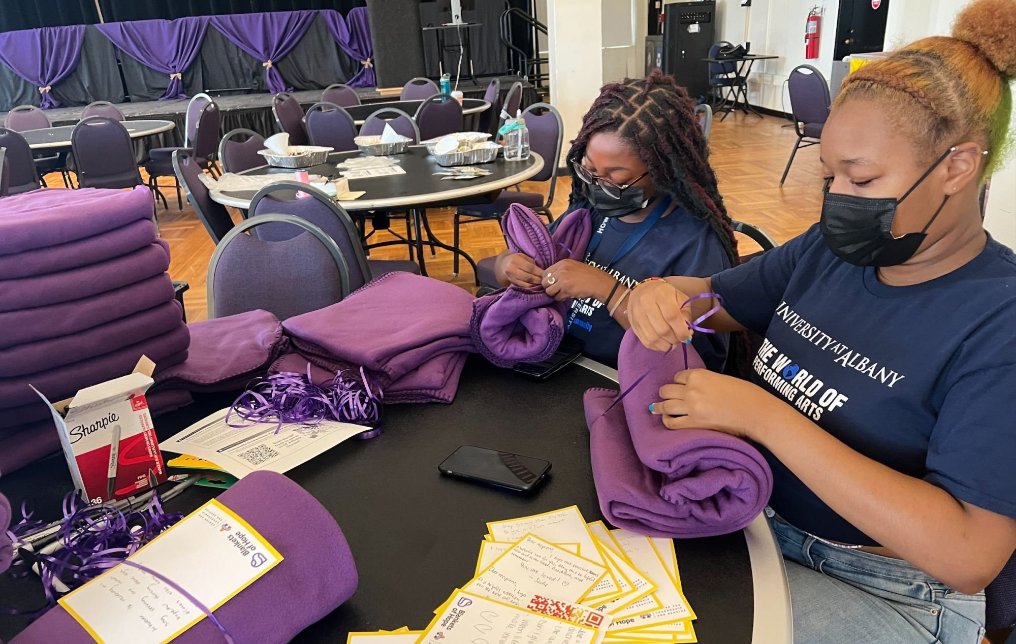 Two masked students in matching navy blue T-shirts that say "The World of Performing Arts" tie ribbons around purple blankets.