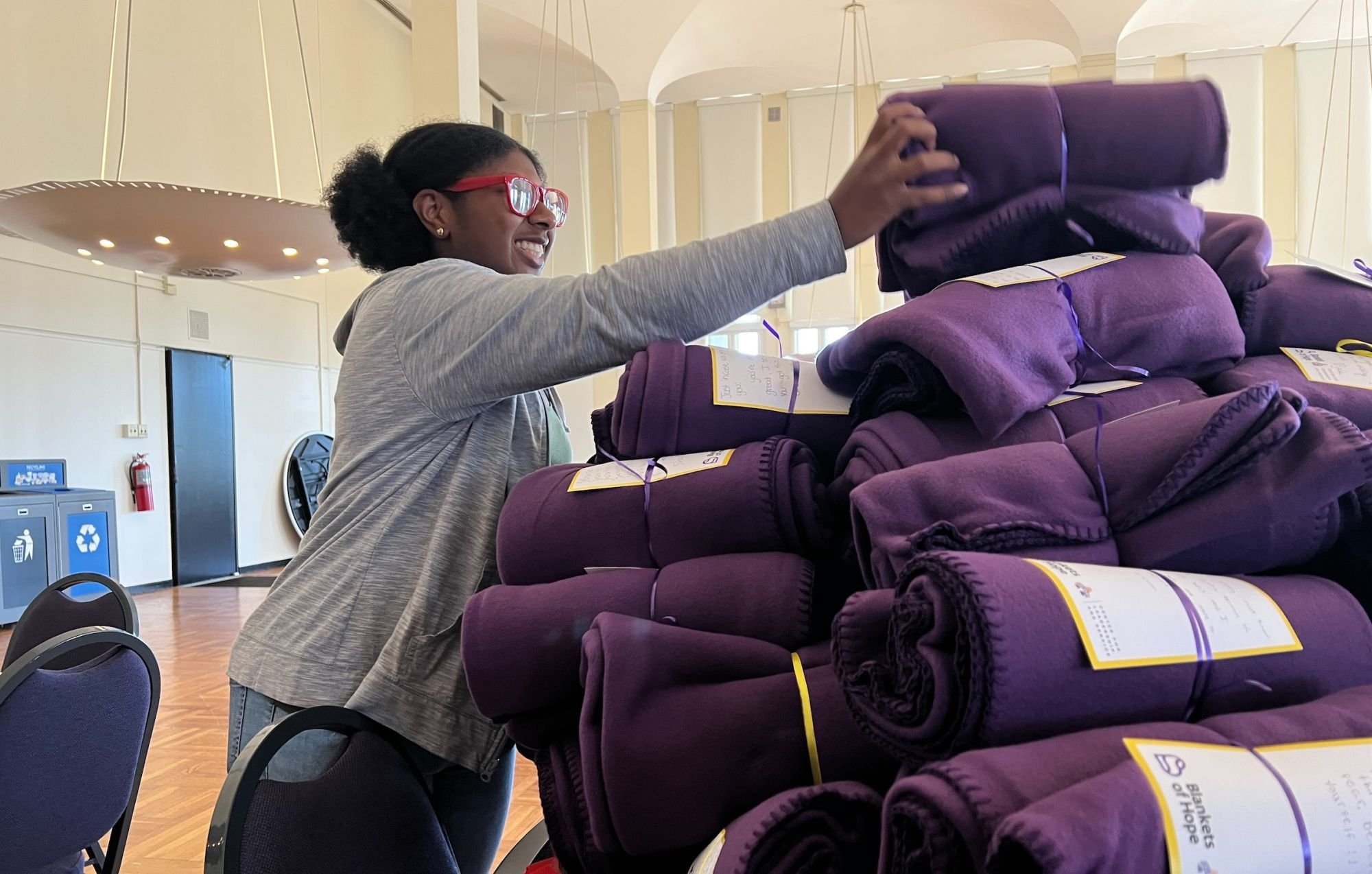 A smiling student places a purple blanket wrapped in ribbon atop a pile of purple blankets destined for area nonprofits.