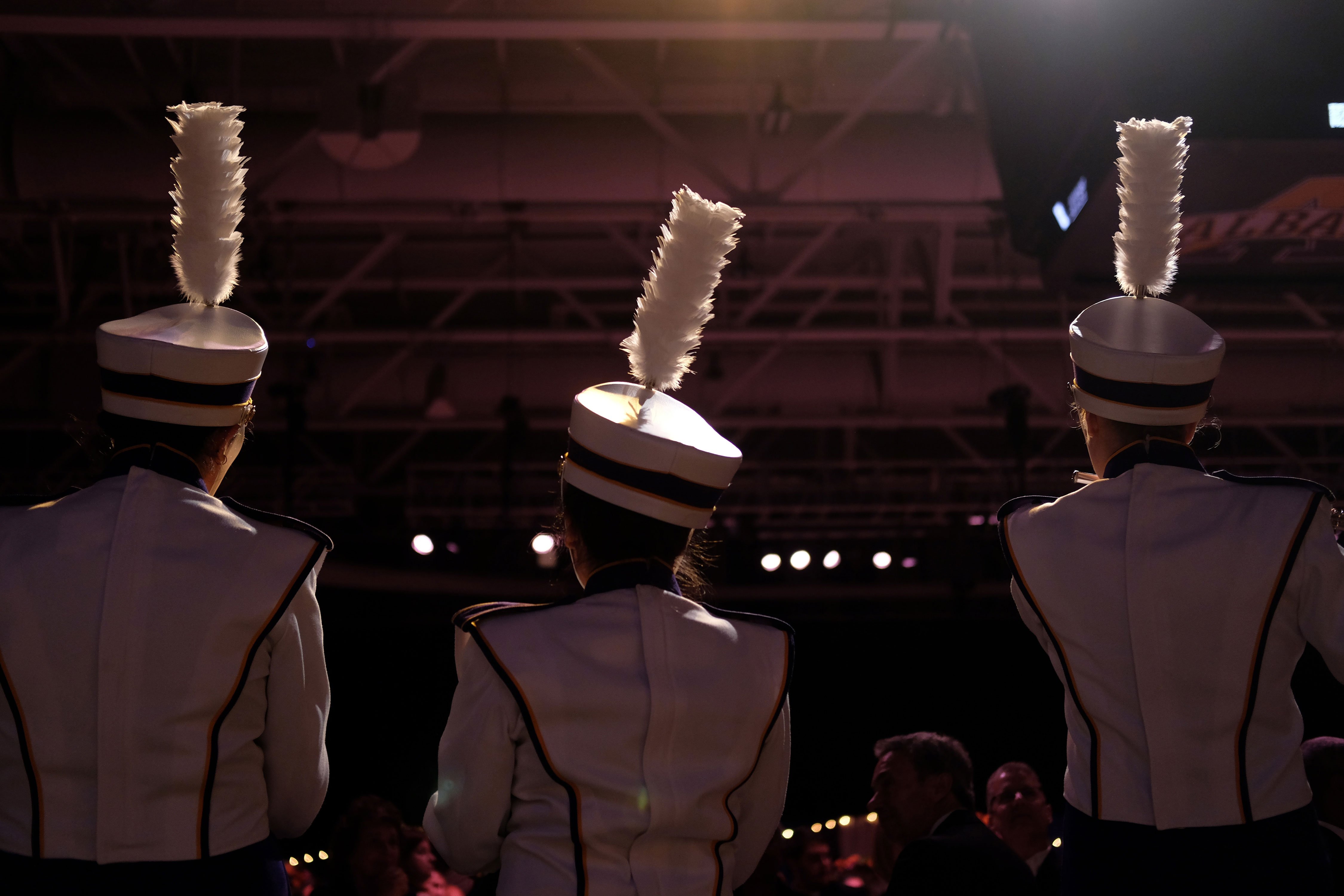 Three students in Marching Band uniforms and hats face away from the camera in a darkened SEFCU Arena.