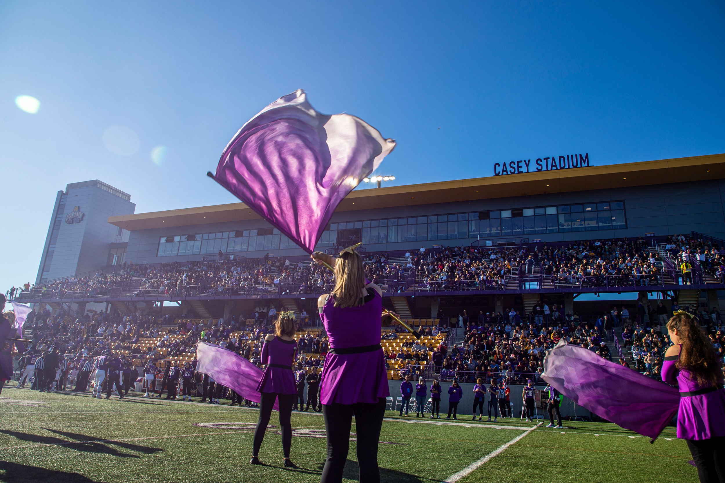 Color guard members wearing purple and black spin purple flags on the field before the packed stands at Casey Stadium on a sunny day.