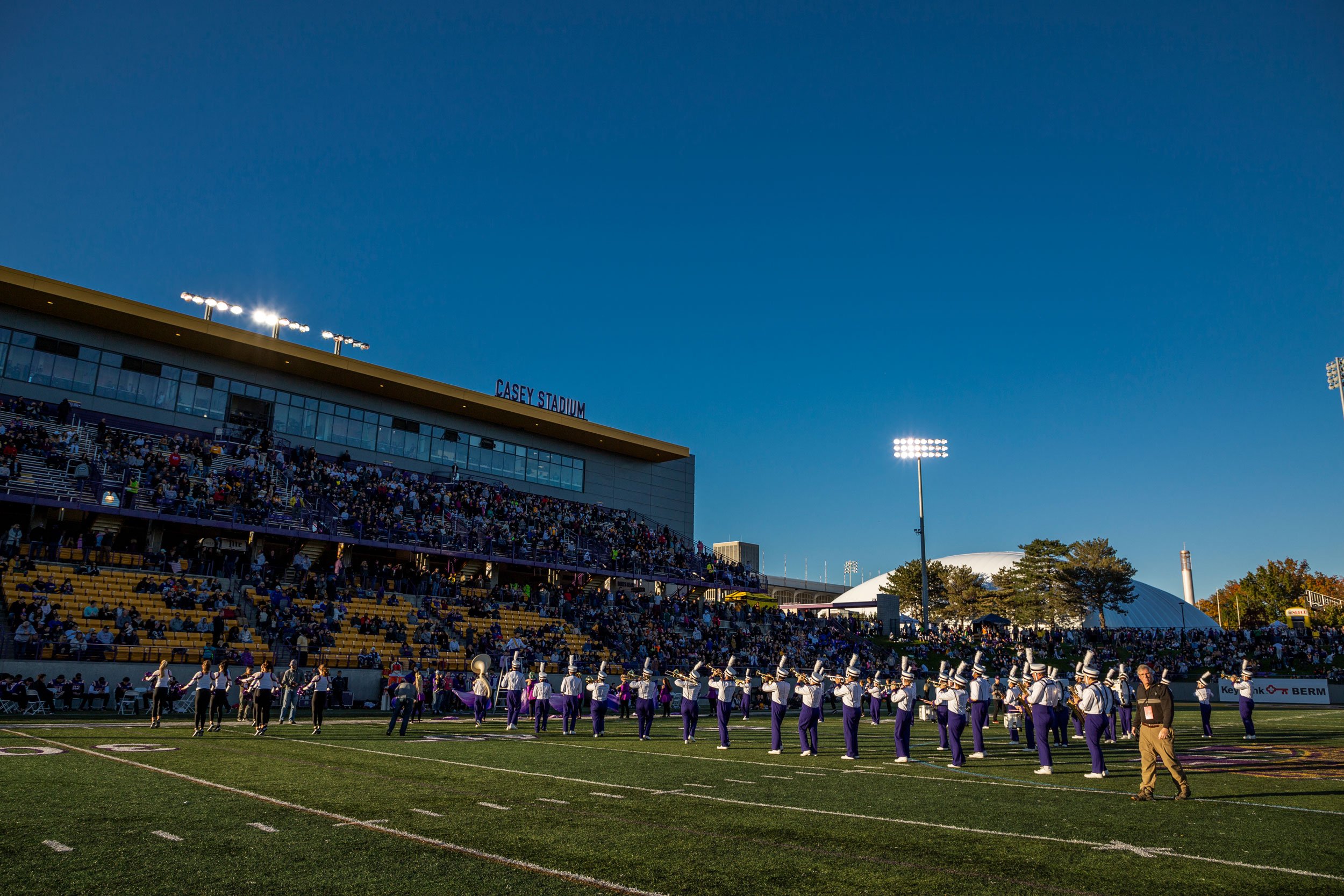 The Marching Great Danes play in formation on the football field before the stands at Casey Stadium under blue skies.