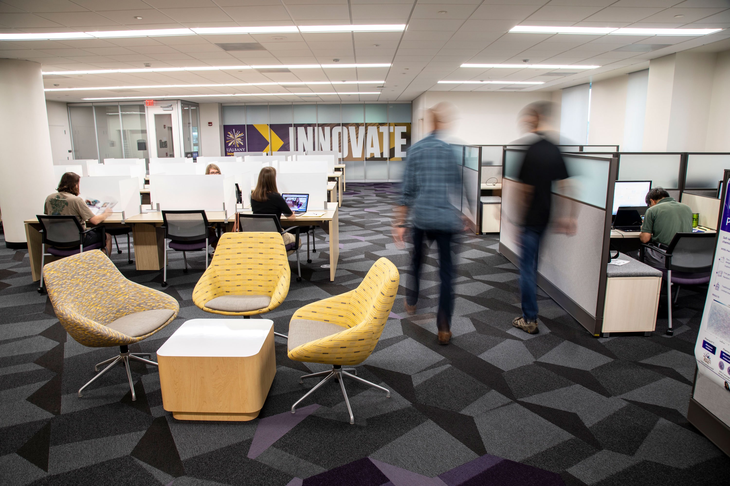Two people are a blur as they walk through the Innovation Center. Others sit at desks around the space. The "innovate" sign is visible in the background.