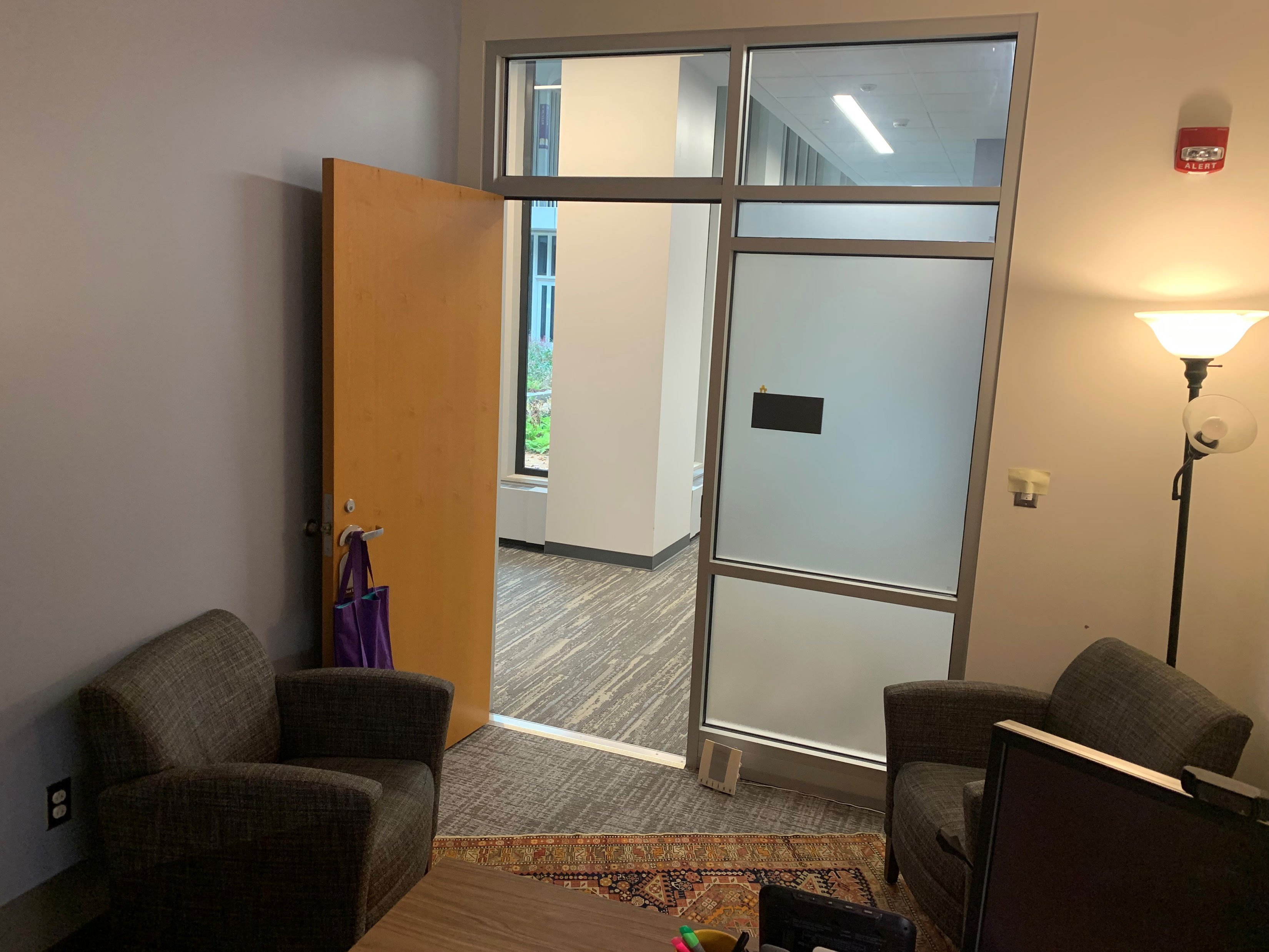 The view of a hallway from inside an office. There are two gray arm chairs and a stand light inside the office, and a door open to a hallway beyond.