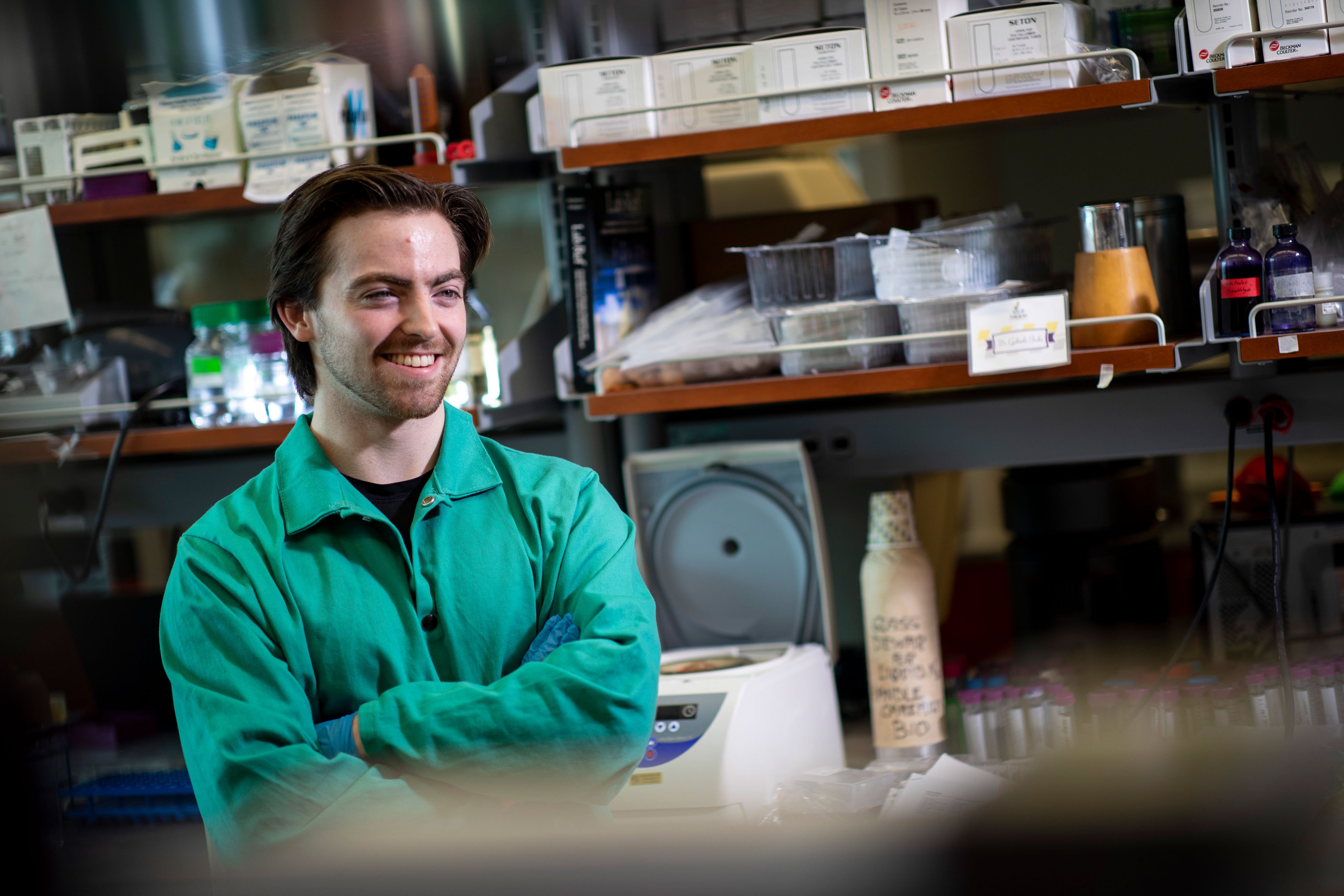 A student in a green lab coat inside a biology laboratory crosses his arms and smiles as he poses for a photo.