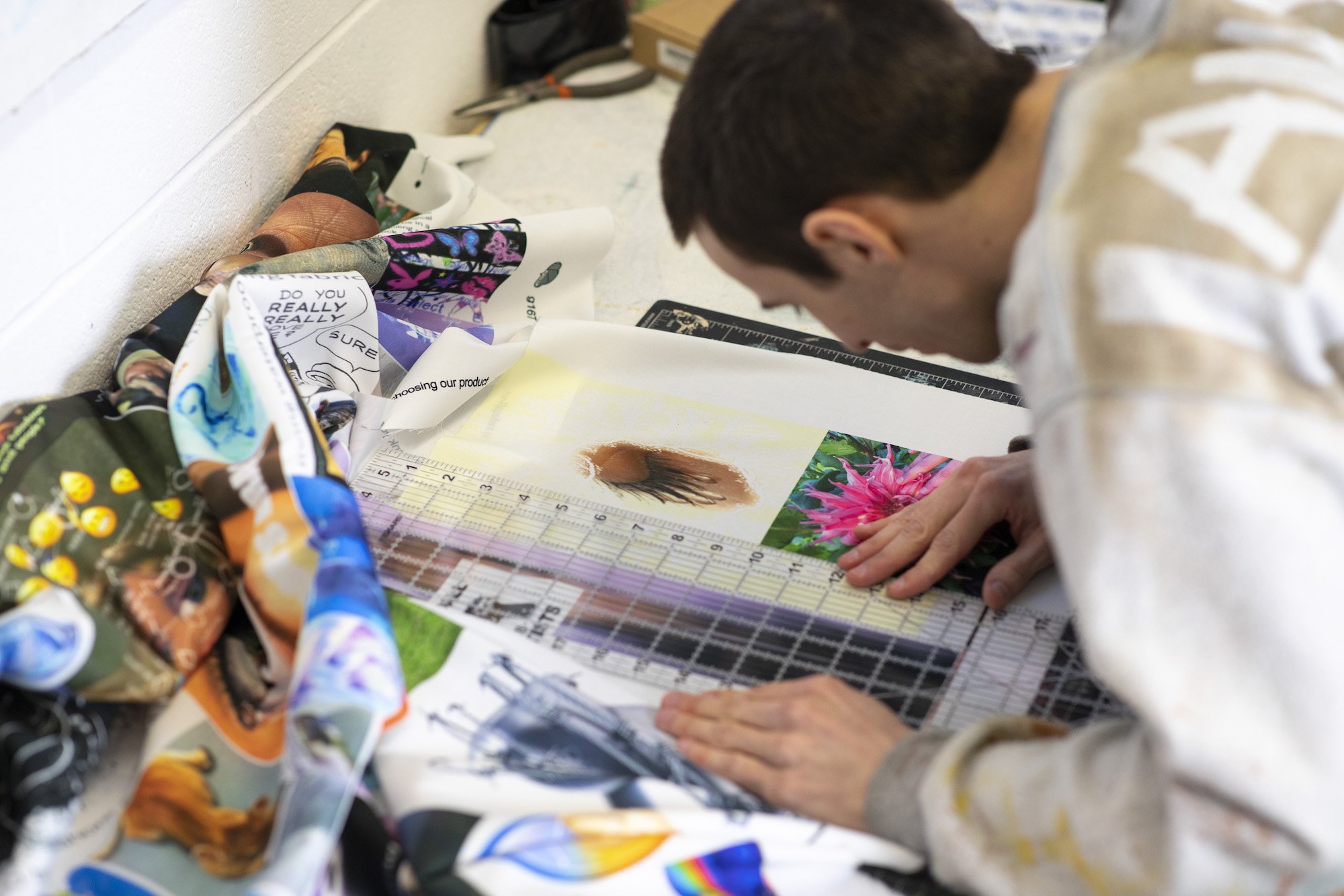 A man with dark hair leans over a desk as he places a ruler next to some pictures surrounded by scraps of fabric.