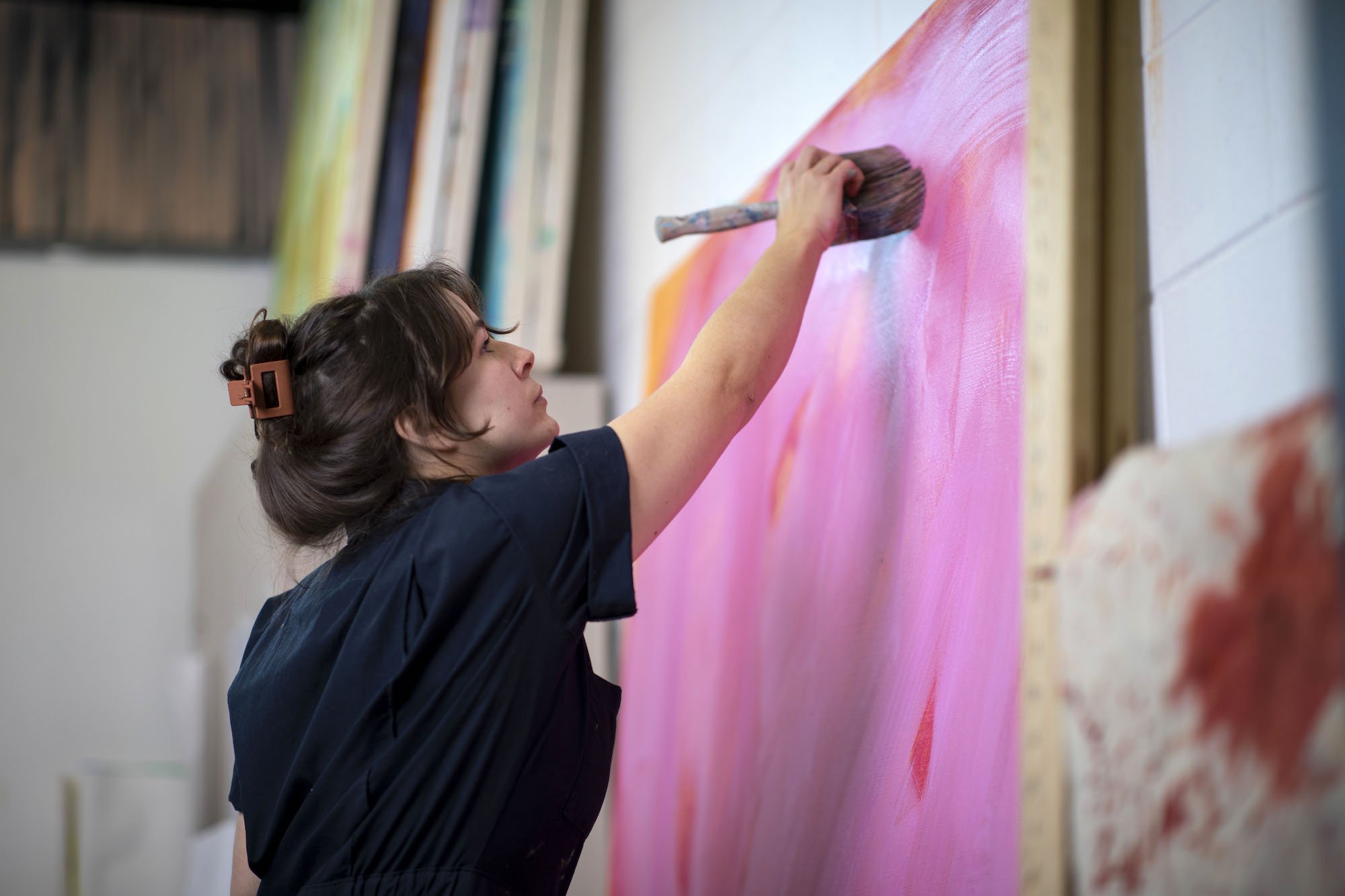 A woman in a navy jumpsuit has an arm extended as she swipes pink paint onto a large canvas frame covered in pink, orange and red paint. 