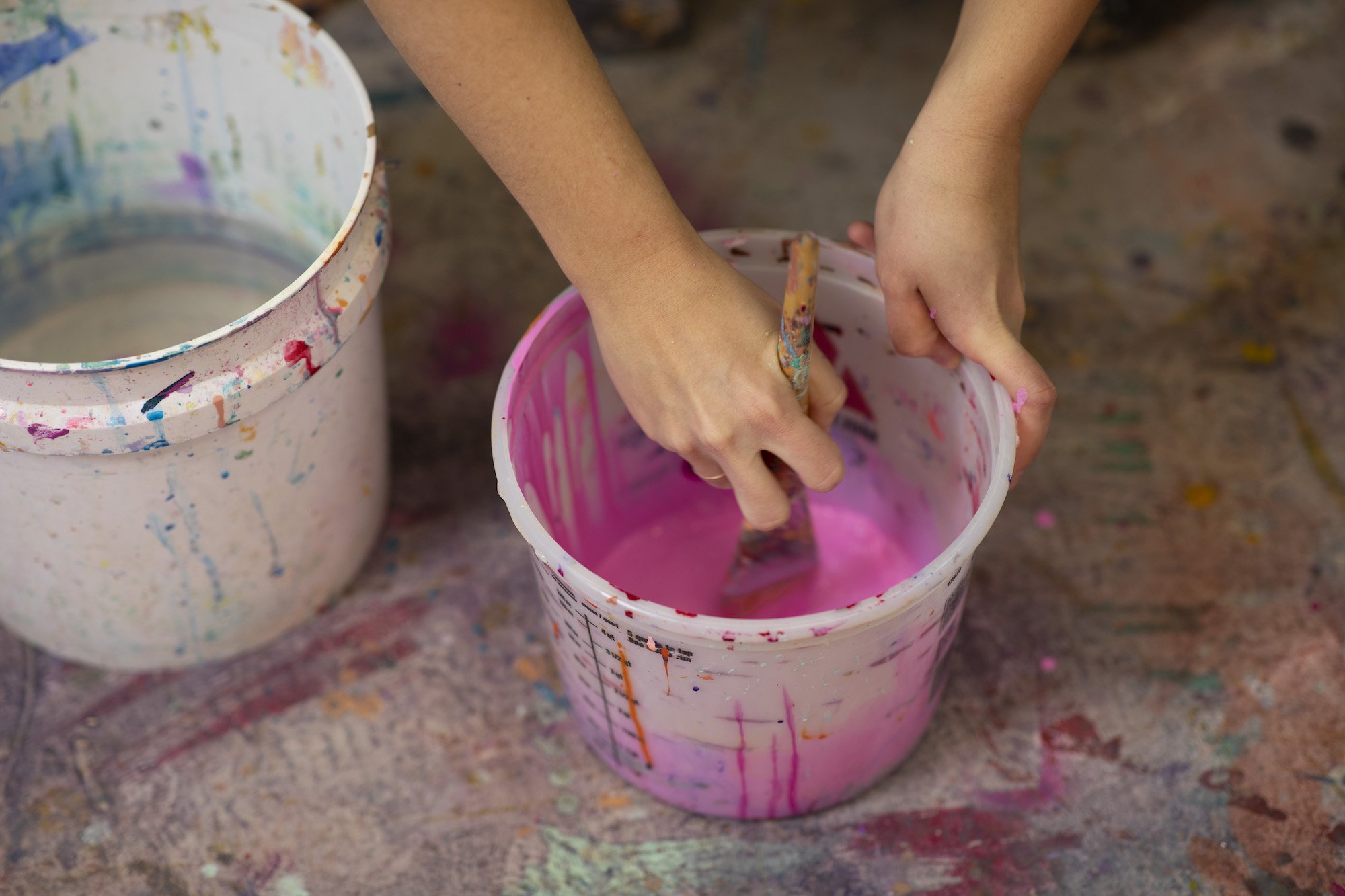 A hand dips a brush into a plastic bucket of pink paint that sits on a paint-splattered concrete floor. A larger bucket holding water sits next to it.