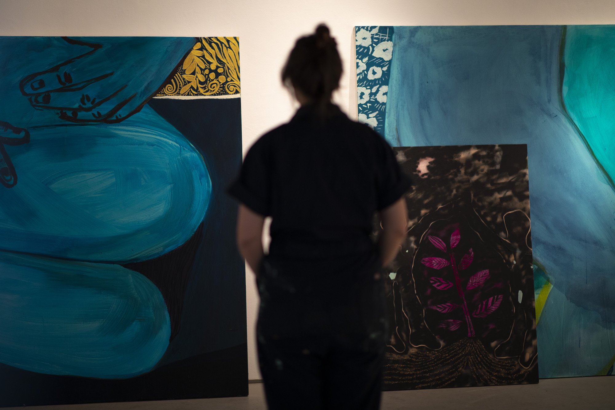 A woman’s silhouette is pictured from behind as she stares at several large paintings depicting figures in blue and a pink tree on a moonlit night. 