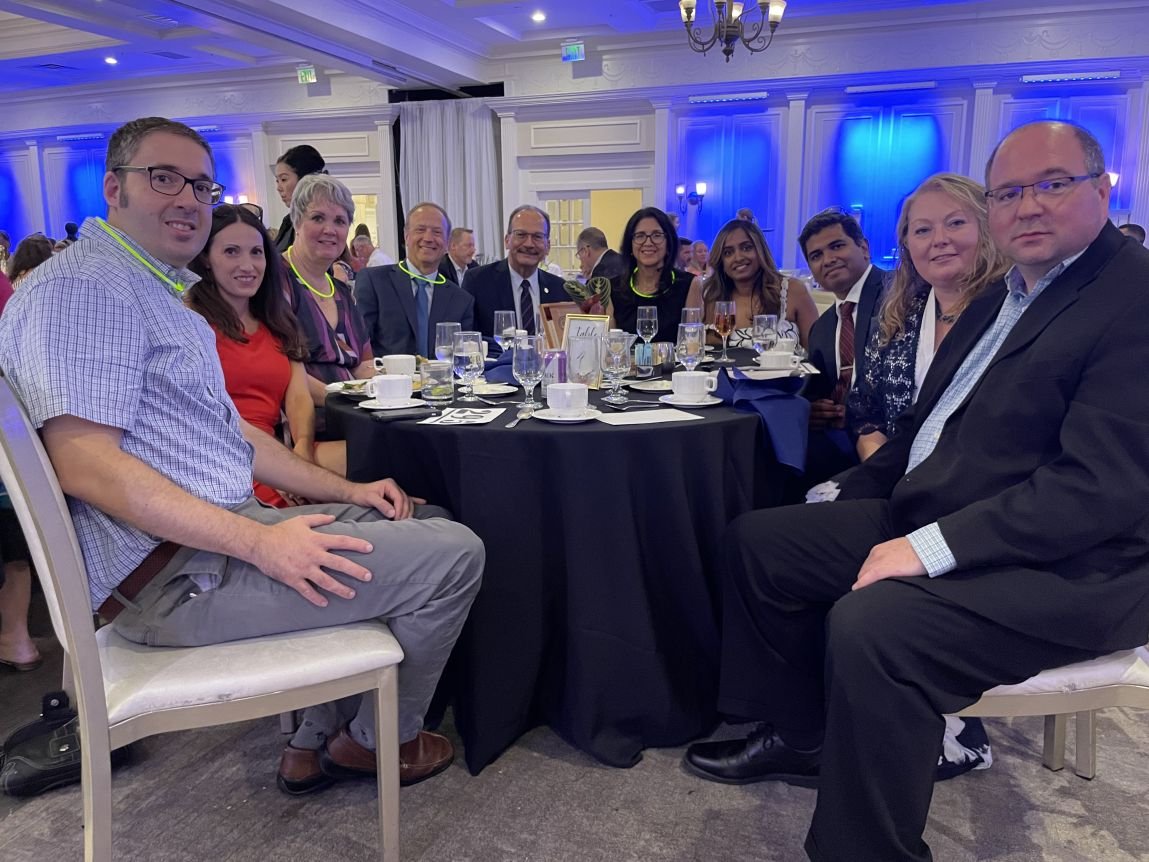  Ten people representing University at Albany leadership and the RNA Institute are seated around a round table for dinner at the Muscular Dystrophy Association event. The background lighting is blue and several people are wearing yellow glow in the dark necklaces. Left to right: Jeremy Logue and his wife Liz Logue, Sheila Seery, Andy Berglund, Havidán Rodríguez and his wife Rosy Rodríguez, Sangeetha Selvam, Jibin Abraham Punnoose, Tammy Reid and John Cleary.