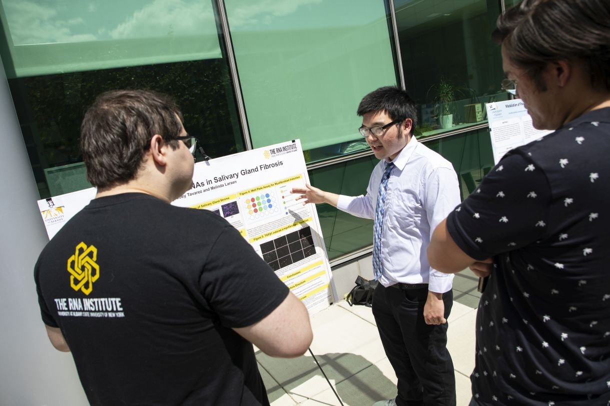A young man wearing a white dress shirt, tie and glasses stands next to his research poster. He is explaining his work to a man wearing a black T-shirt that says 'RNA Institute' on the back. 