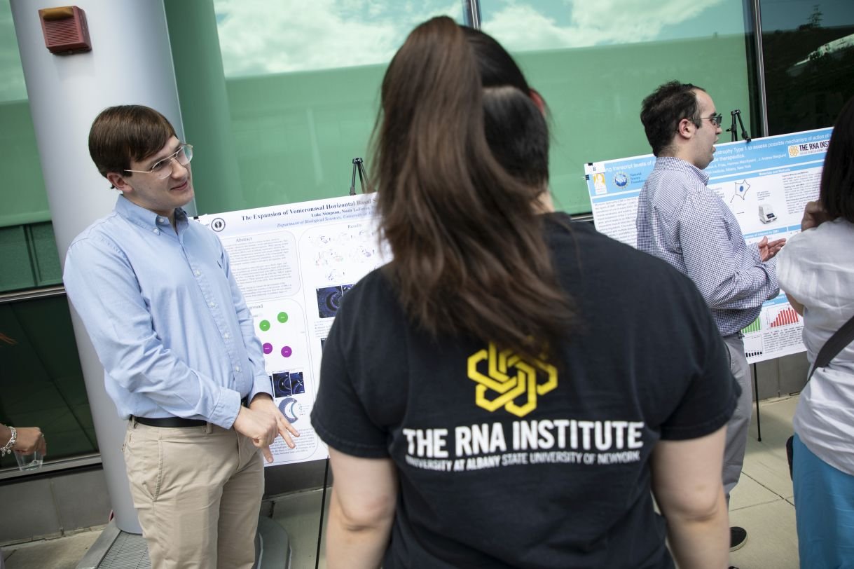 A young man wearing a light blue dress shirt and glasses stands, smiling, next to his research poster. He is explaining his work to a woman wearing a black shirt that says 'RNA Institute' on the back. 