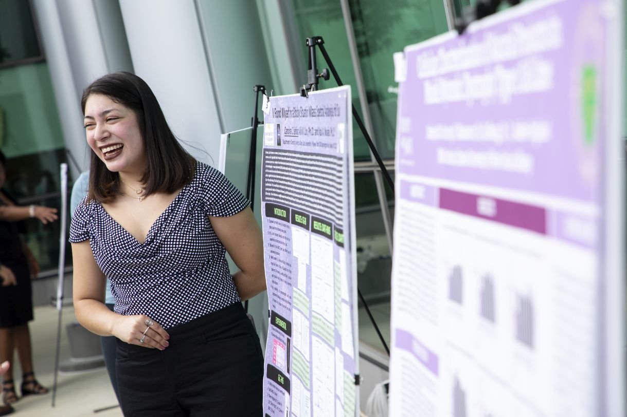 A young woman with short dark hair stands, smiling, next to her research poster. The photo was taken outdoors in the life sciences building courtyard on a sunny day. 