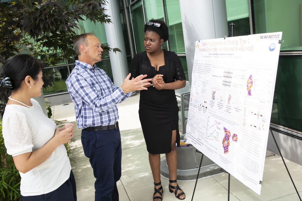 A young woman wearing a black dress and black heeled sandals speaks with a man wearing a blue plaid dress shirt. The young woman is an REU student and she is presenting her research poster at the RNA Institute's RNA Day celebration. 