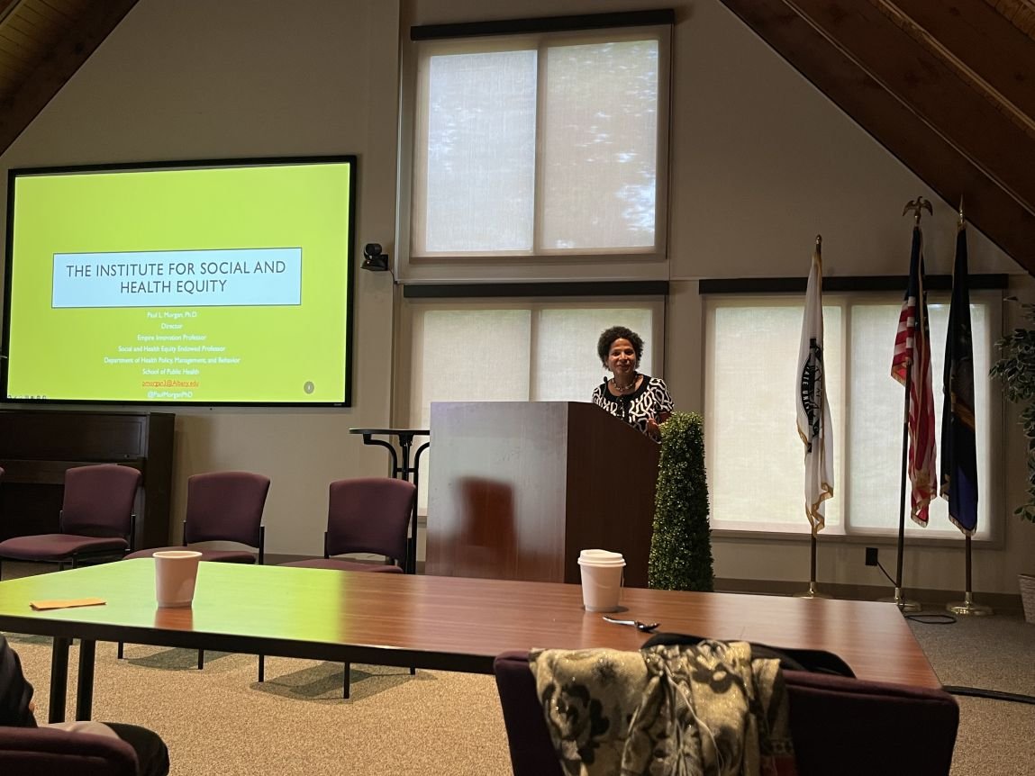 Elizabeth Vásquez stands at a podium delivering a presentation. A lime green PowerPoint side to her left reads: “The Institute for Social and Health Equity” 
