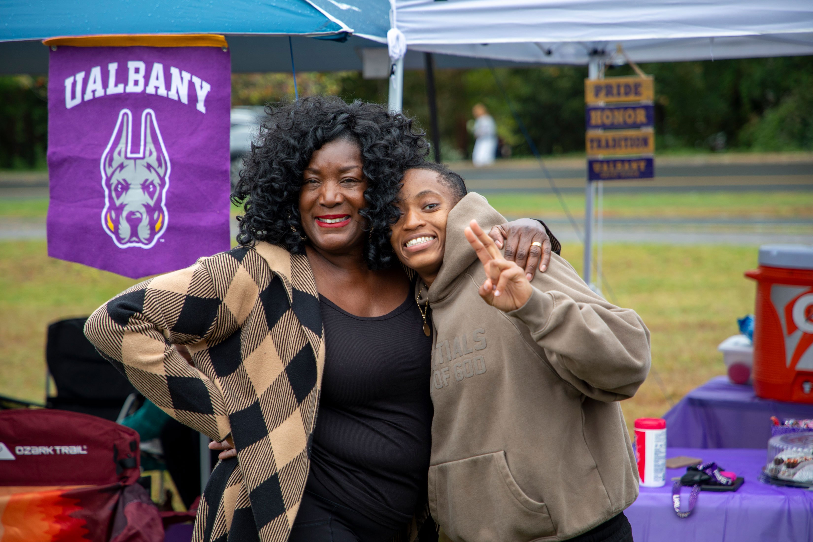 Two adults smile as they pose for a photo in front of a UAlbany sign.