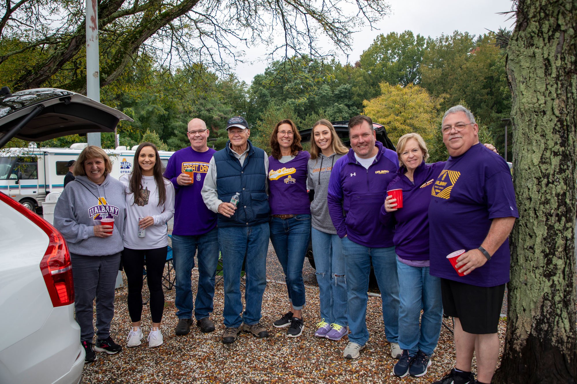 Nine adults, most of whom are wearing UAlbany gear, smile as they pose for a photo while tailgating.