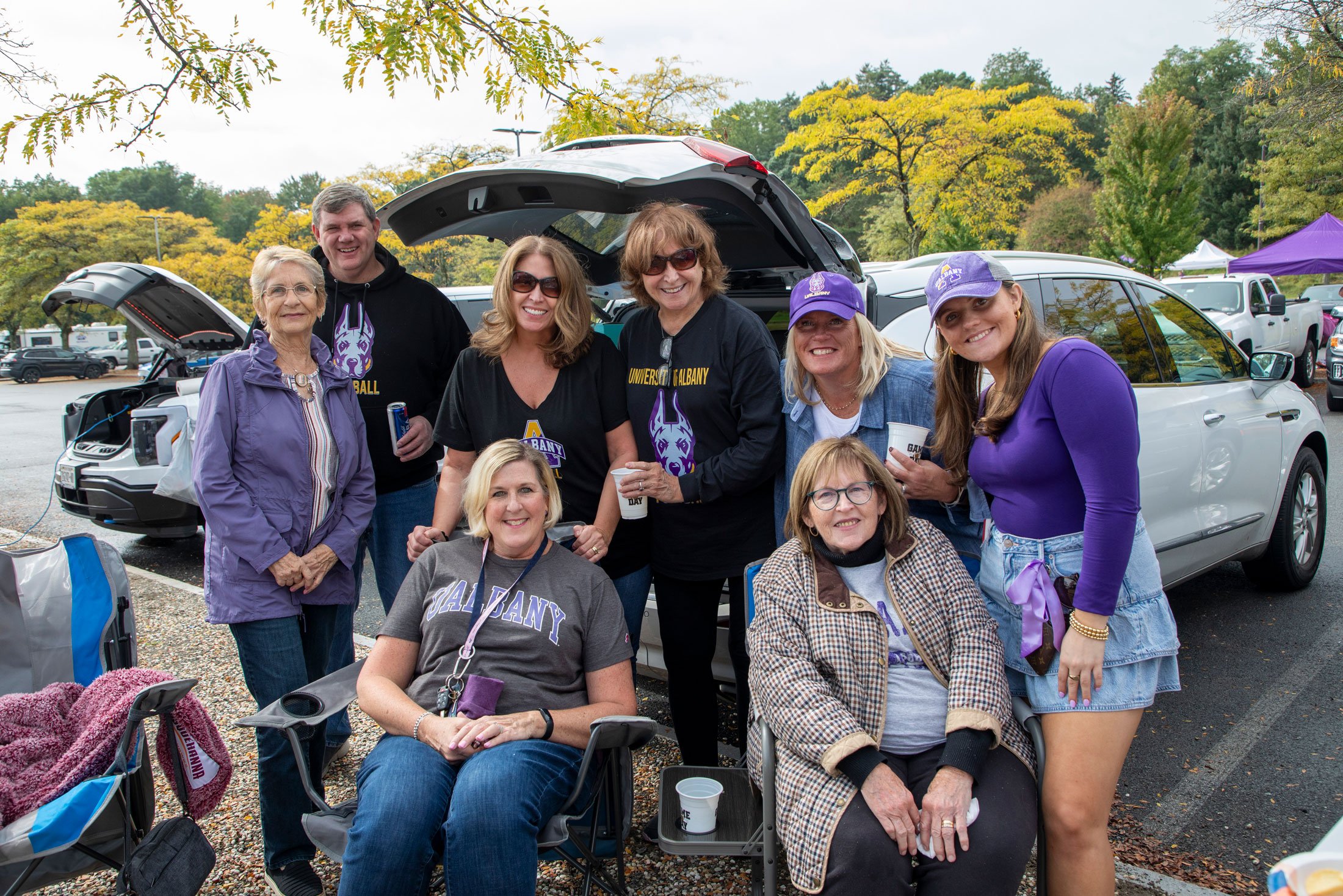 Eight adults, two of whom are sitting in camp chairs, smile as they pose for a photo while tailgating.